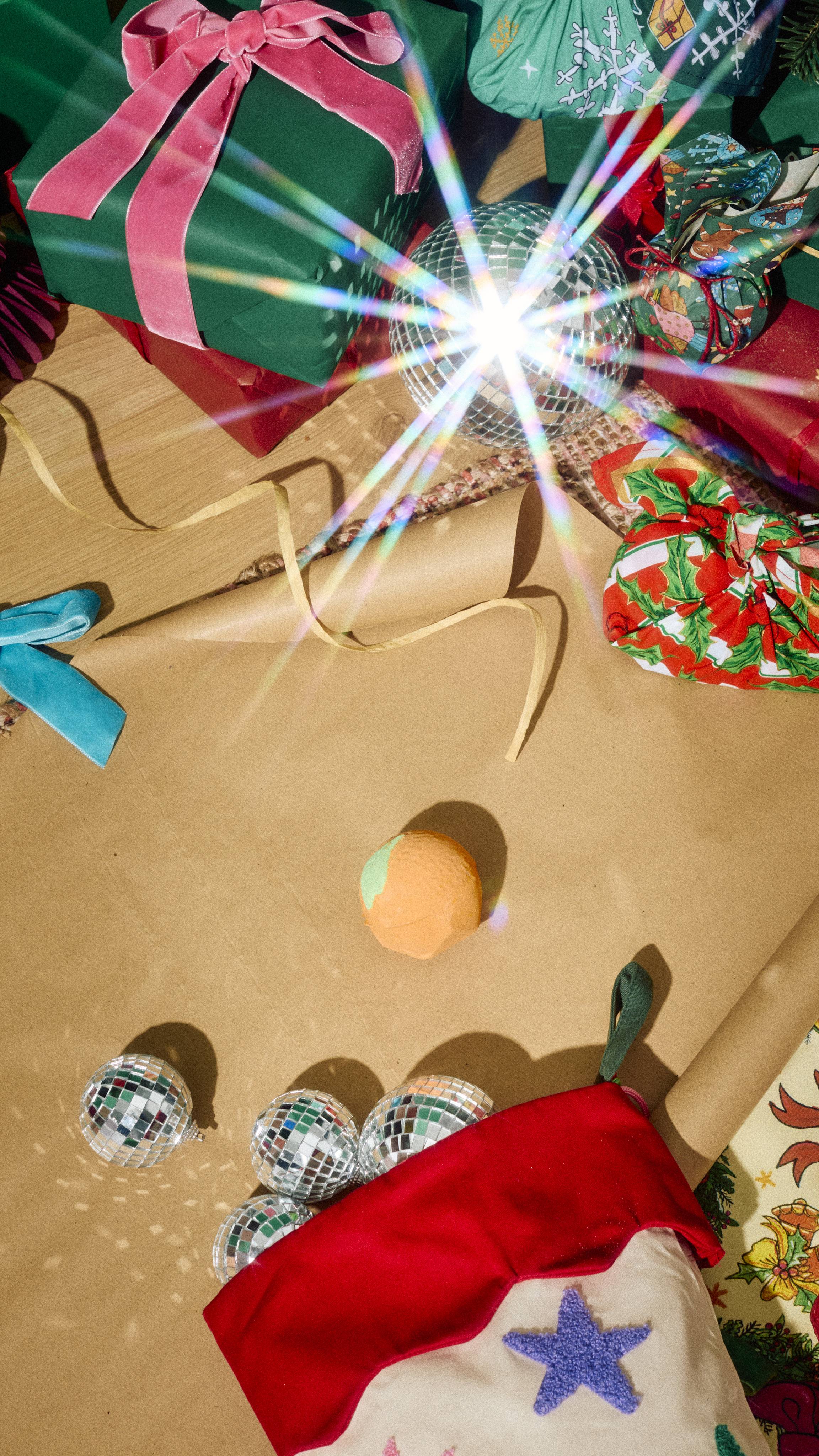 An orange bath bomb sits on brown paper alongside mini disco balls in a stocking, wrapped gifts with pink velvet ribbon, and colourful knot-wraps with sparkling light effects.