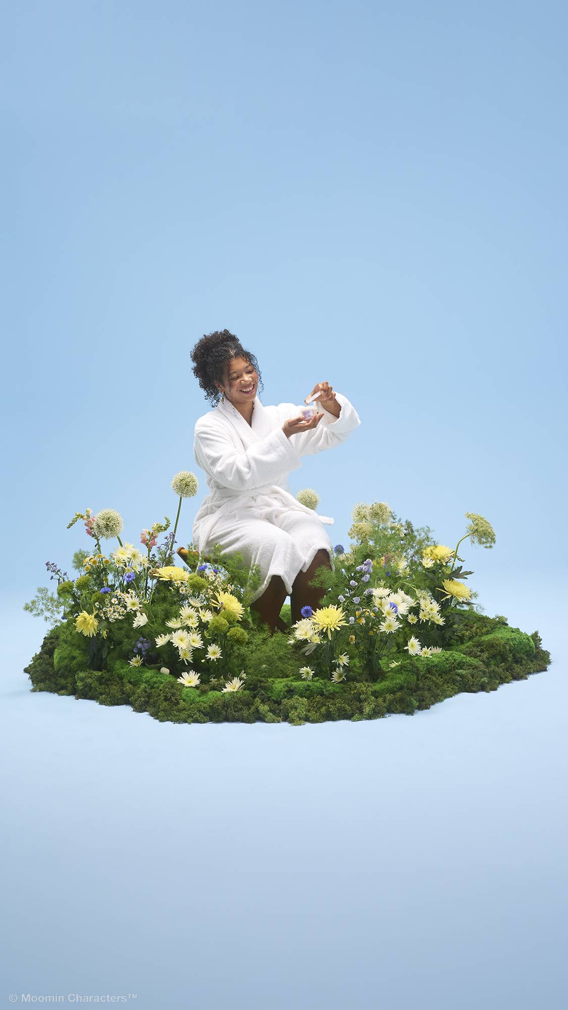 A smiling person wearing a white dressing gown sits amongst a lush patch of green moss and vibrant yellow and white wildflowers. They are holding open a small tub of Moominmamma hand and body lotion, all set against a solid light blue background.