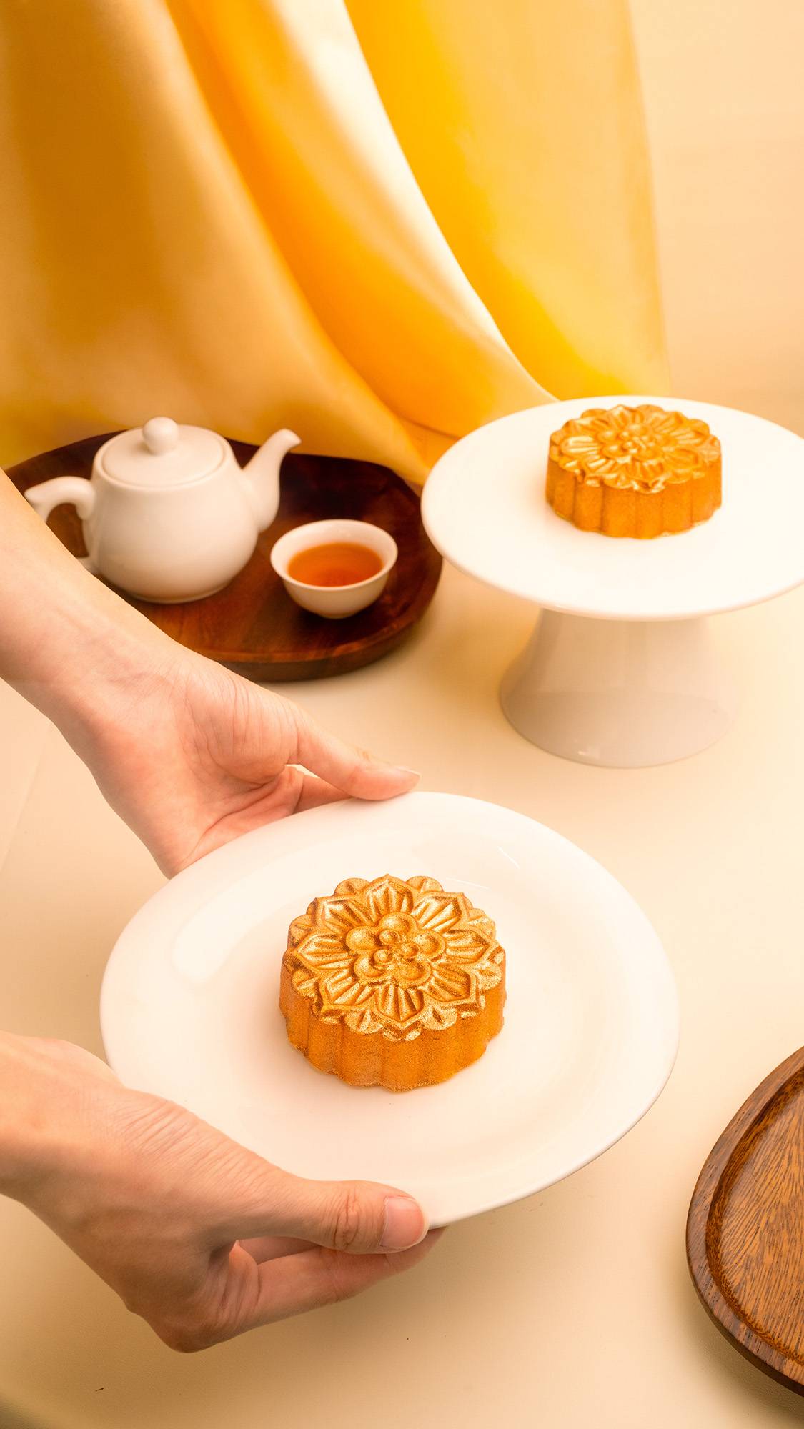 A pair of hands presents a golden, intricately patterned Lush "Mooncake" bath bomb on a white plate. In the background, another mooncake sits on a white stand next to a teapot and teacup on a wooden tray, set against a warm yellow drape.