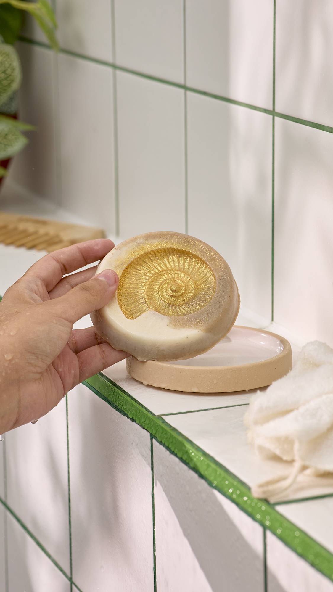 A hand holding a round cream-coloured Lush Ms Ammonite soap bar embossed with a golden shimmering ammonite shell design, held above a ceramic soap dish against tiled bathroom walls.   