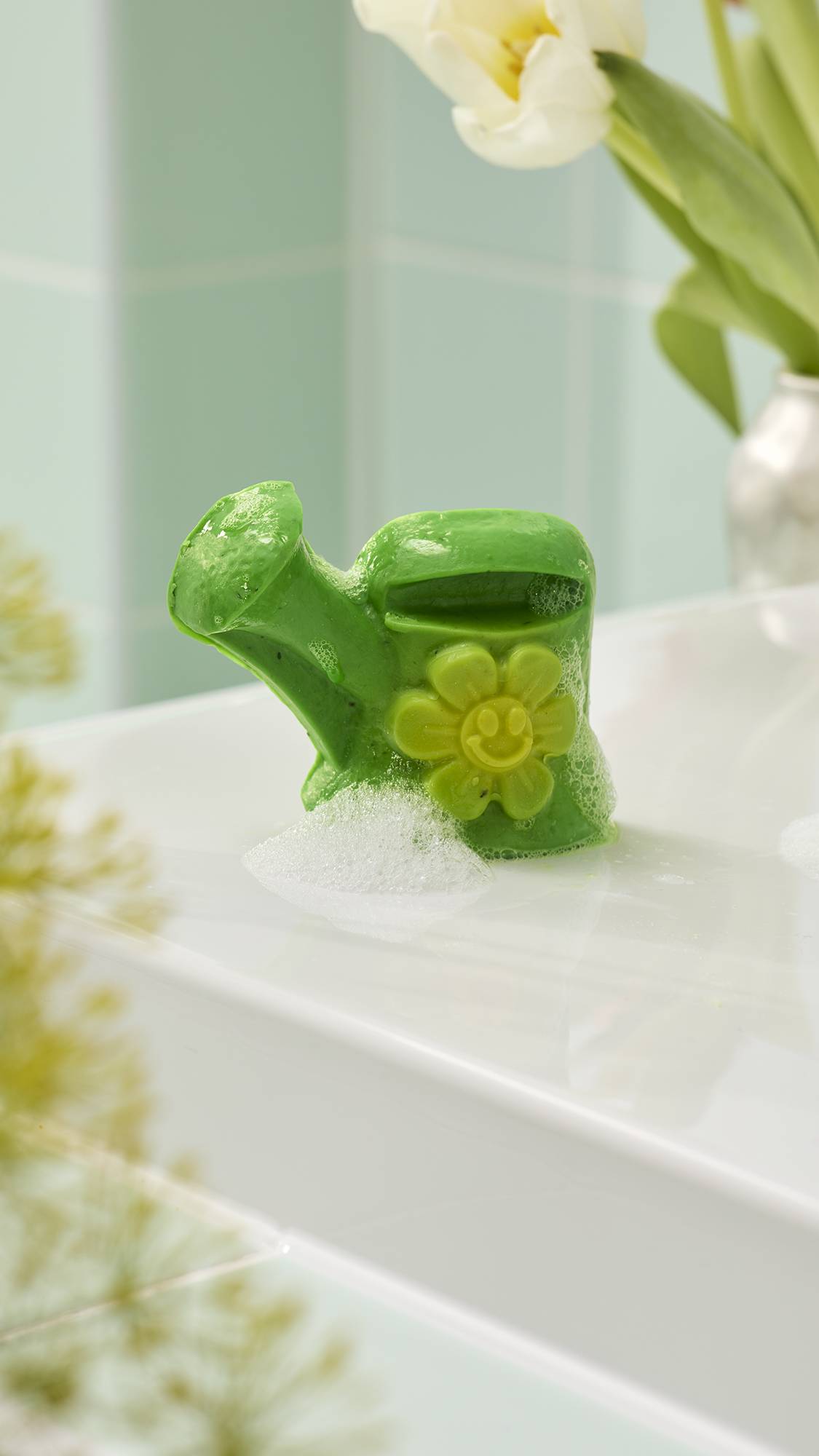 A wet, green "Mum's Garden" soap shaped like a watering can sits on a white surface, featuring a yellow smiley-face flower. White foam gathers at the base, with blurred white tulips and green tiles in the background.