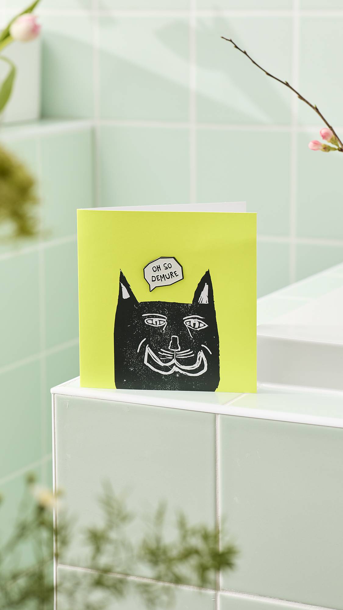A square, lime-green Oh So Demure greetings card featuring a whimsical illustration of a black cat's head. A white speech bubble above the cat reads: "OH SO DEMURE" in black capital letters. The speech bubble is also a pin badge.