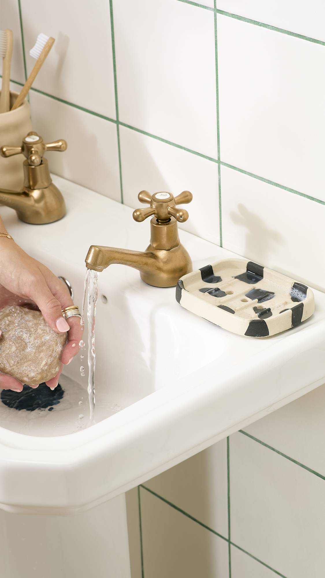 A person holds a bar of soap under running water from a brass tap over a white bathroom sink, with a cream and black cow-print soap dish resting beside the tap against white tiled walls.   