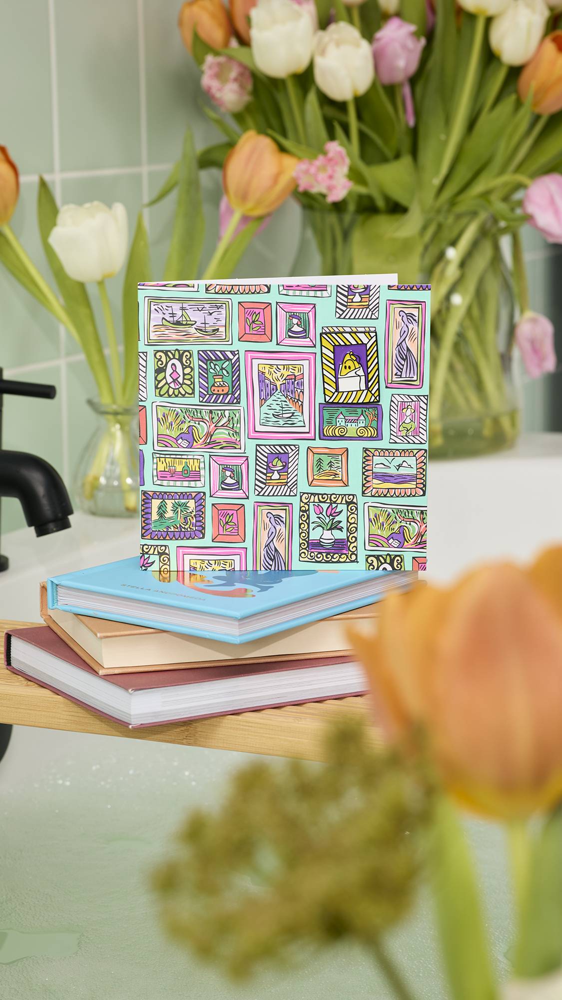 An illustrated "Pictures For You: greetings card sits on a stack of books atop a wooden bath tray. The card features a colourful, hand-drawn gallery wall pattern on a teal background with a detachable enamel pin badge.