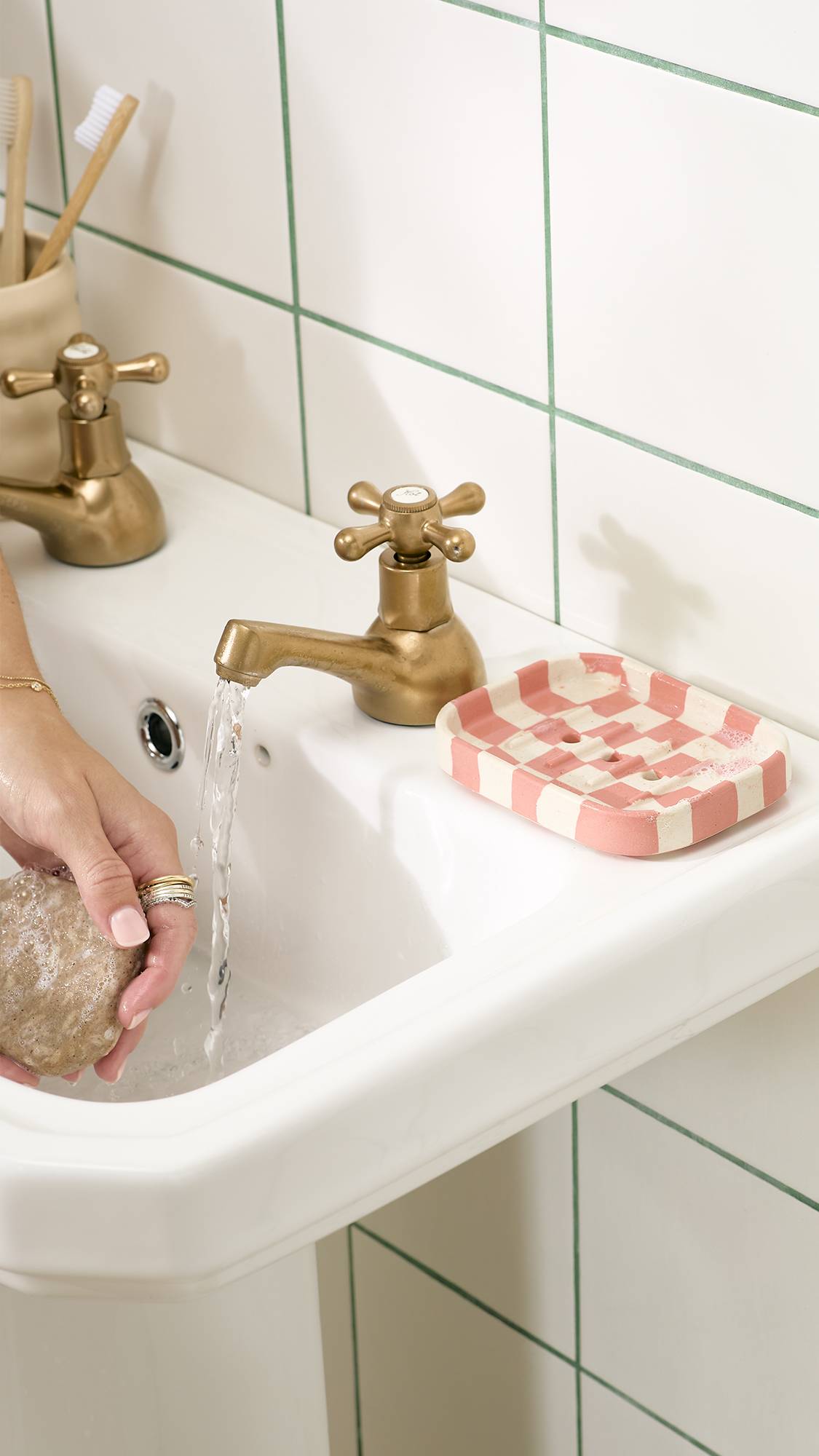 A person holds a bar of soap under running water from a brass tap over a white bathroom sink, with a pink and cream chequered ceramic soap dish resting beside the tap against white tiled walls.   
