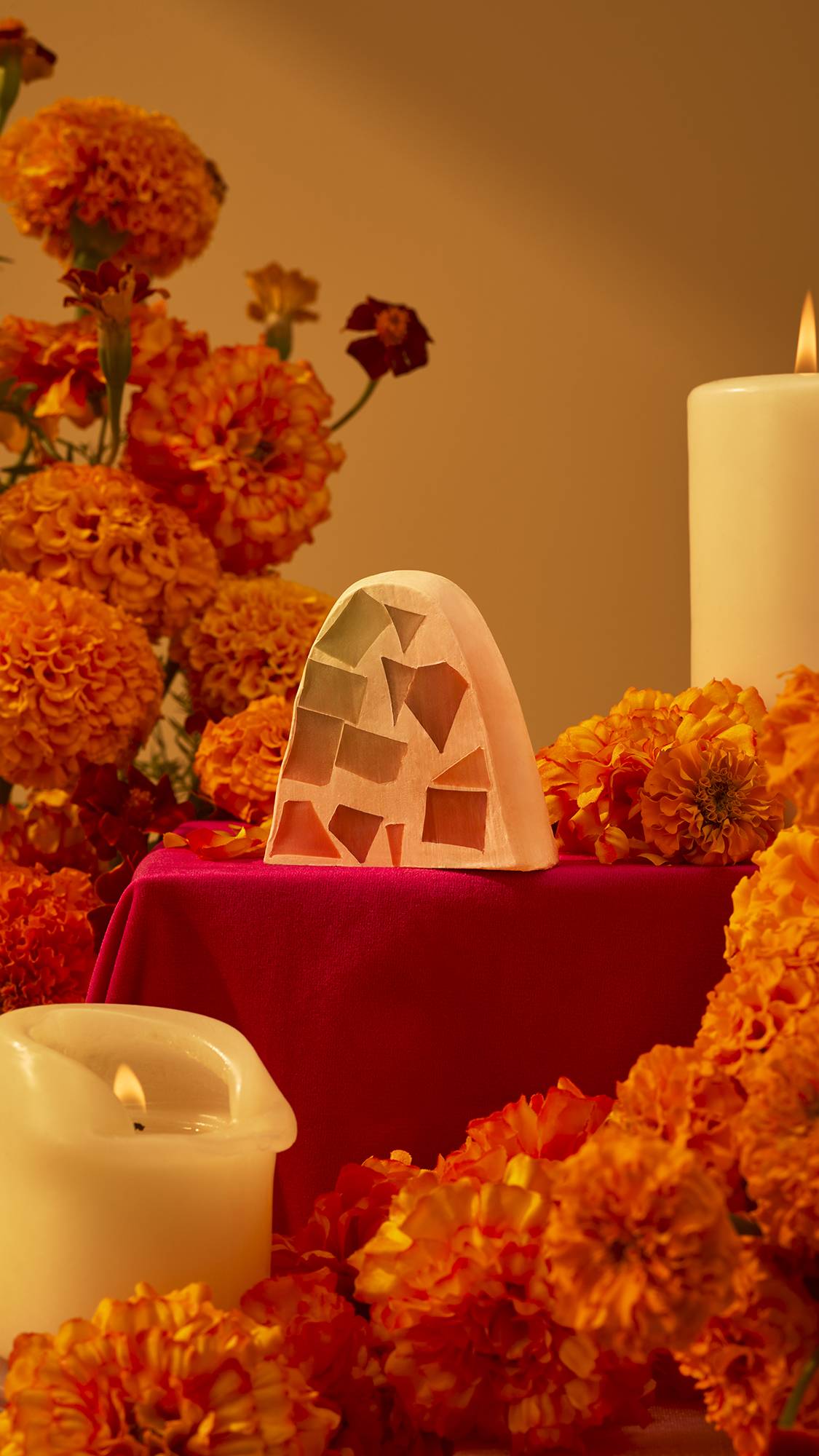 An arch-shaped mosaic bar of Postrecito soap rests on a red draped pedestal, surrounded by vibrant orange marigolds and lit candles in a warm setting.
