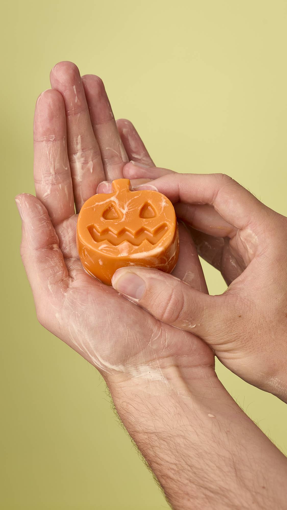 A person holds a wet Lush pumpkin facial gummy in their hands, which are lightly lathered with a white foam. The carved jack-o'-lantern face is visible on the orange bar against a plain, light olive-green background.
