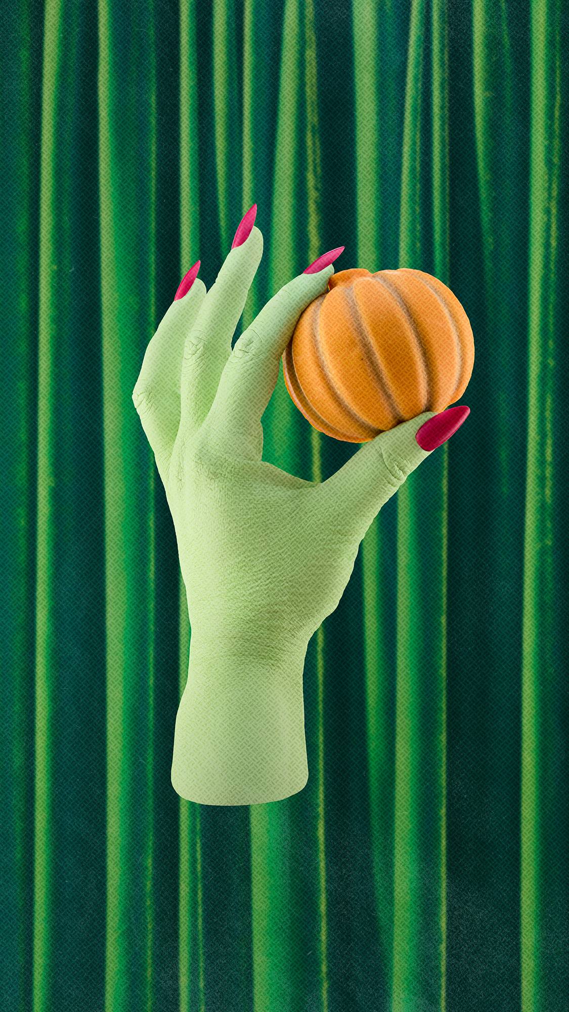 A green hand with long, bright pink nails holds an orange pumpkin-shaped solid body scrub between its fingers, set against a background of draped green velvet curtains.