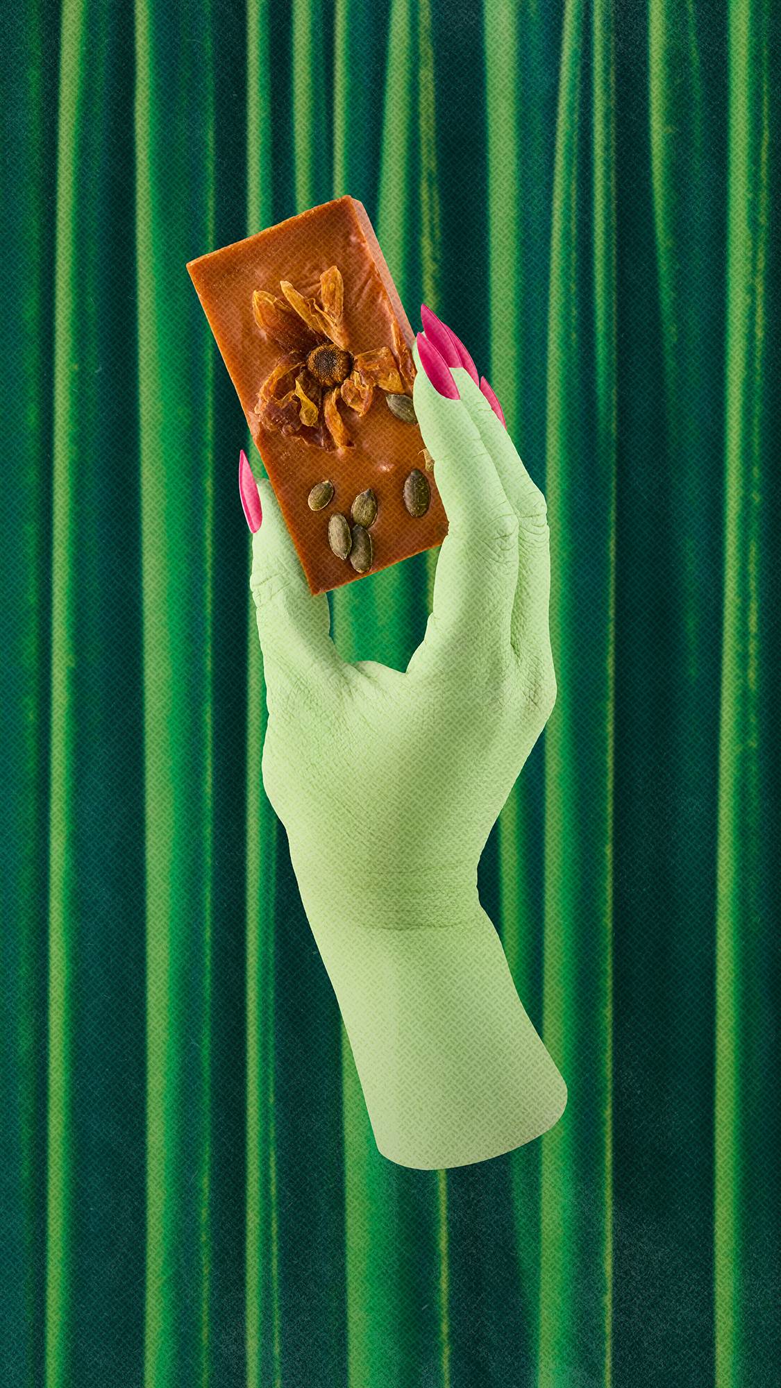 A green-skinned hand with bright pink nails holds an orange "Pumpkin Spice" soap topped with a dried flower and seeds, set against rich, dark green velvet curtains.