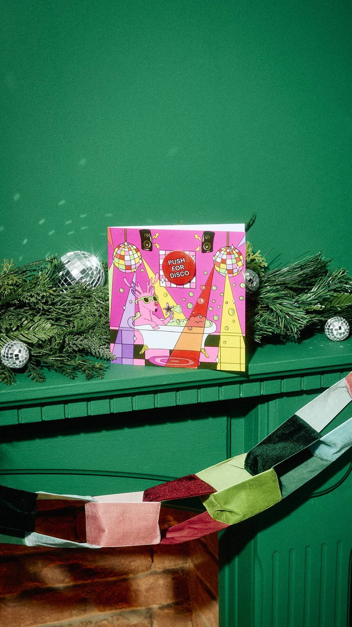 A vibrant pink Christmas card illustrating a reindeer in a bathtub with disco lights and a red badge reading "Push for Disco" sits on a green mantelpiece surrounded by pine garland, mini disco balls, and a fabric chain.