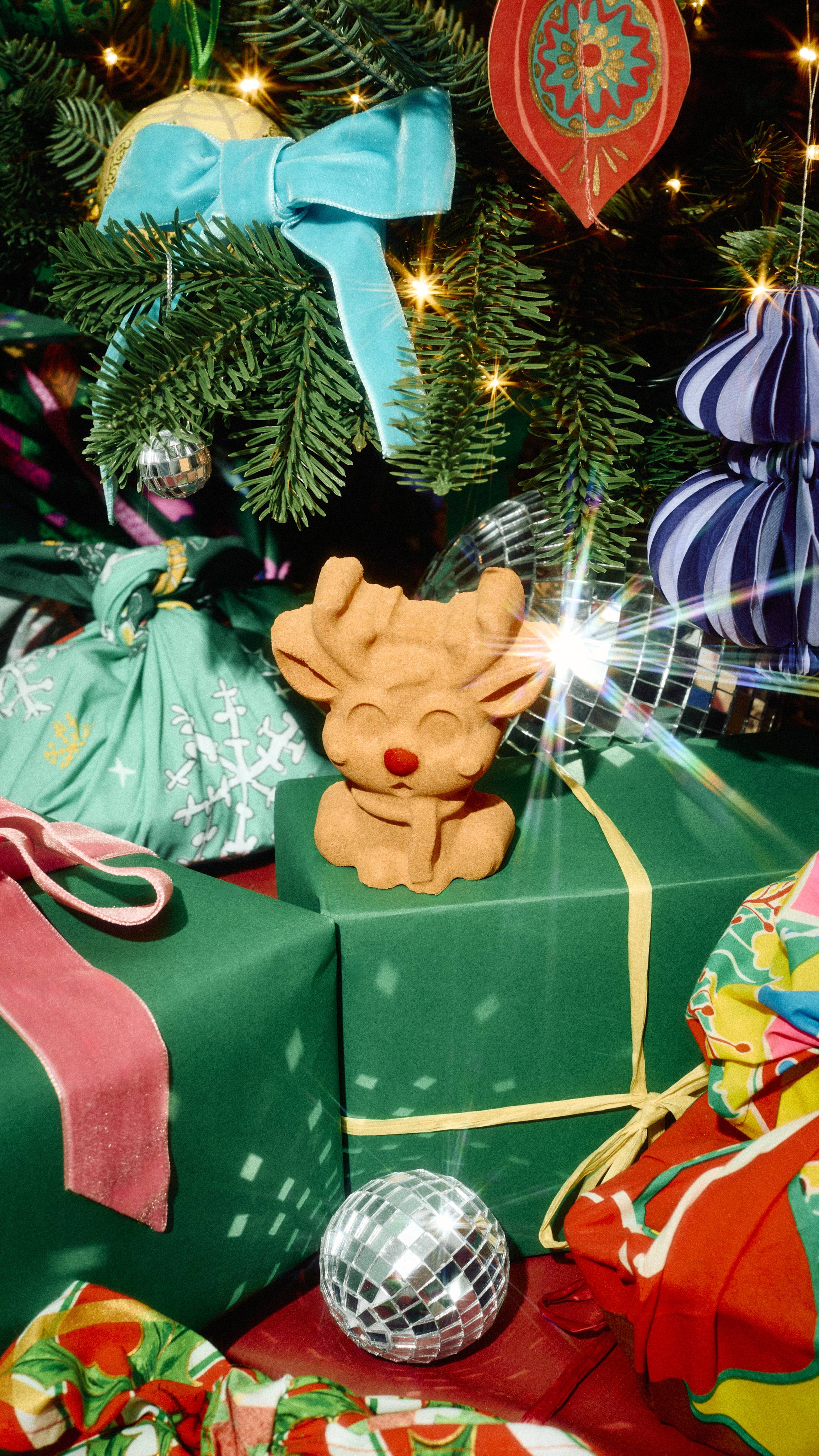 A light brown reindeer-shaped bath bomb sits on a green wrapped present under a Christmas tree decorated with a blue velvet bow, sparkling baubles, and festive ornaments.