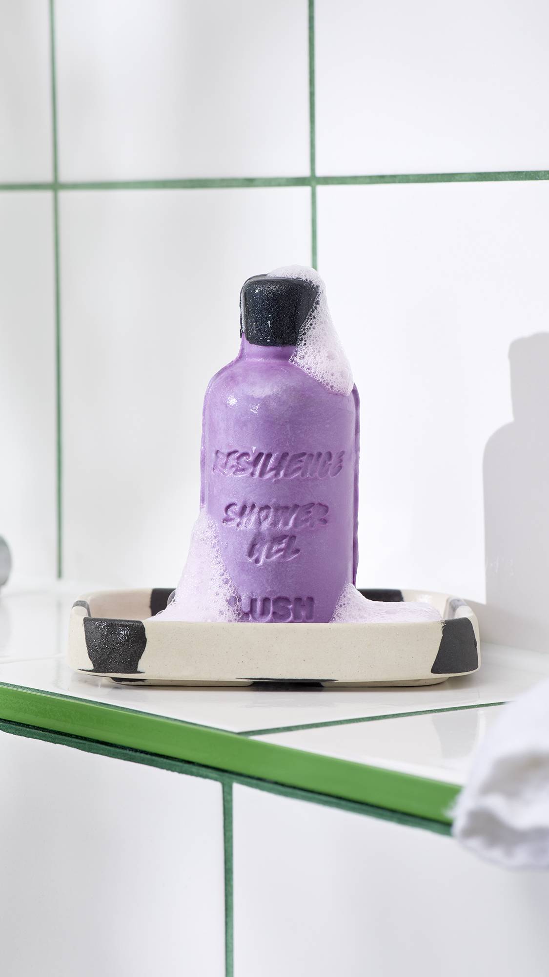 A bright purple solid shower gel shaped like a bottle rests on a black and cream spotted soap dish, partially covered in frothy white foam. It sits against a background of white tiles with green grouting.