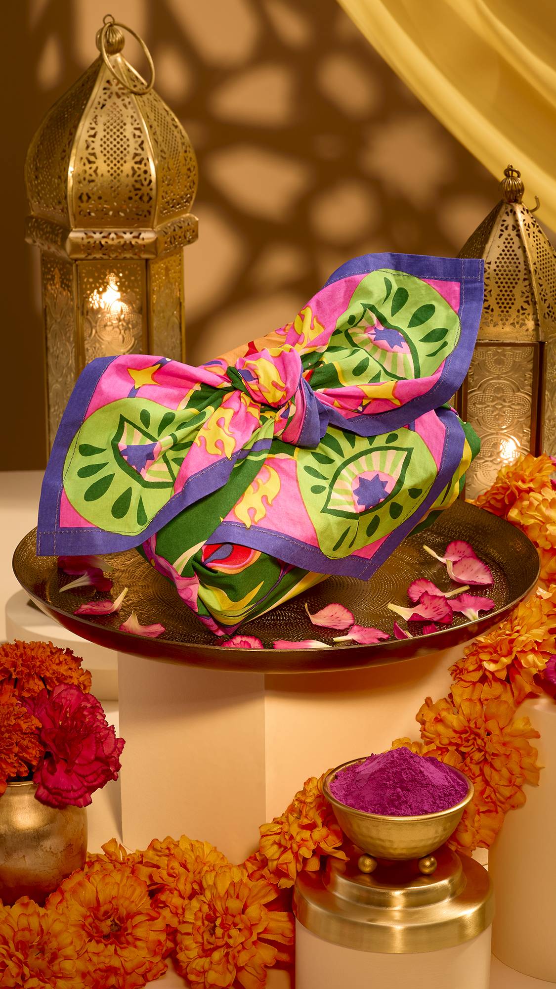 A gift wrapped in a vibrant green, pink, and purple patterned Lush Knot Wrap rests on a decorative tray with rose petals, surrounded by marigolds, purple pigment, and lit brass lanterns.