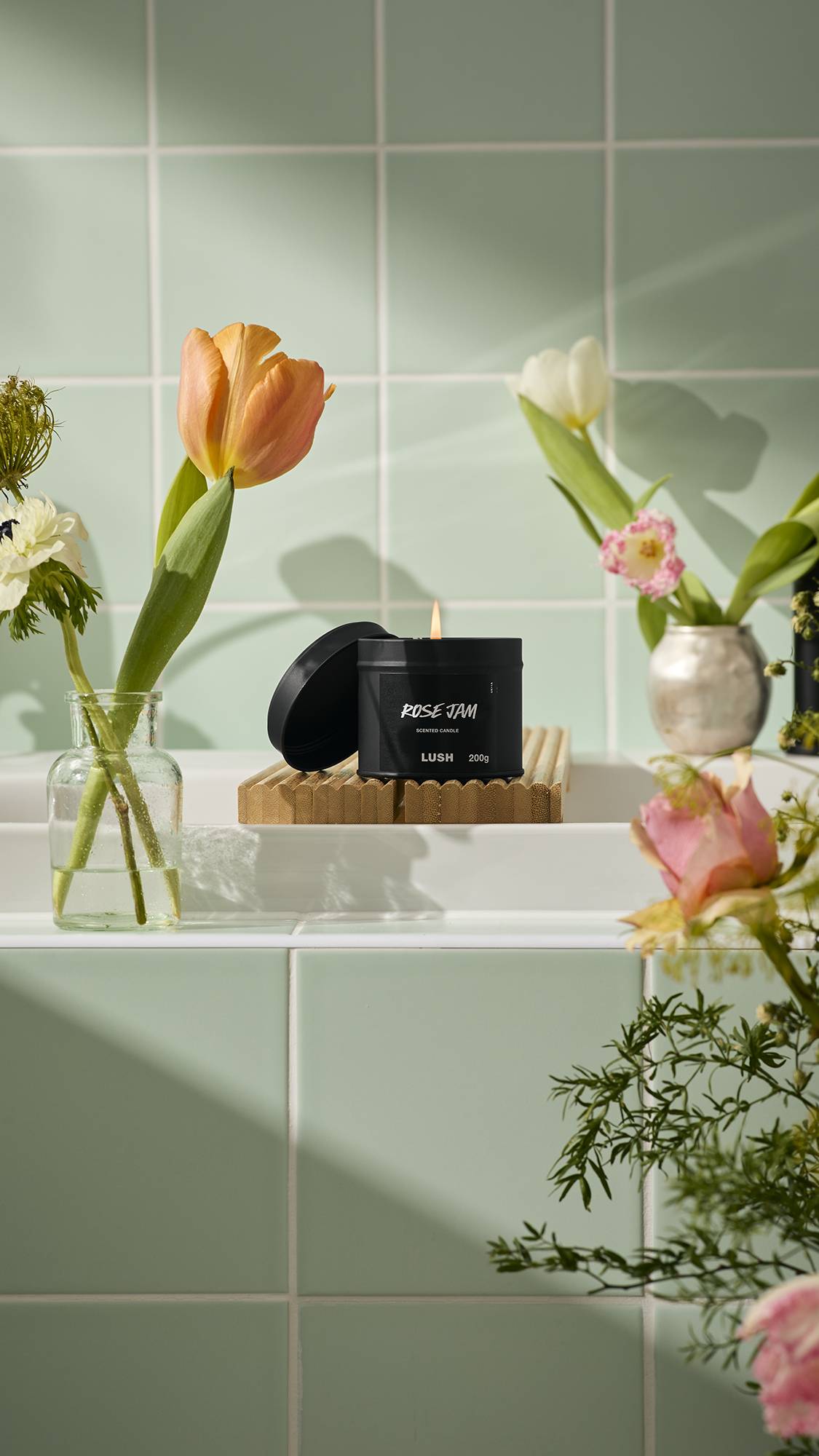 The image shows a round, black tin of Lush "Rose Jam" scented candle sitting lit on a wooden bath bridge. It is surrounded by vases of orange and white tulips against a backdrop of pale green tiles.
