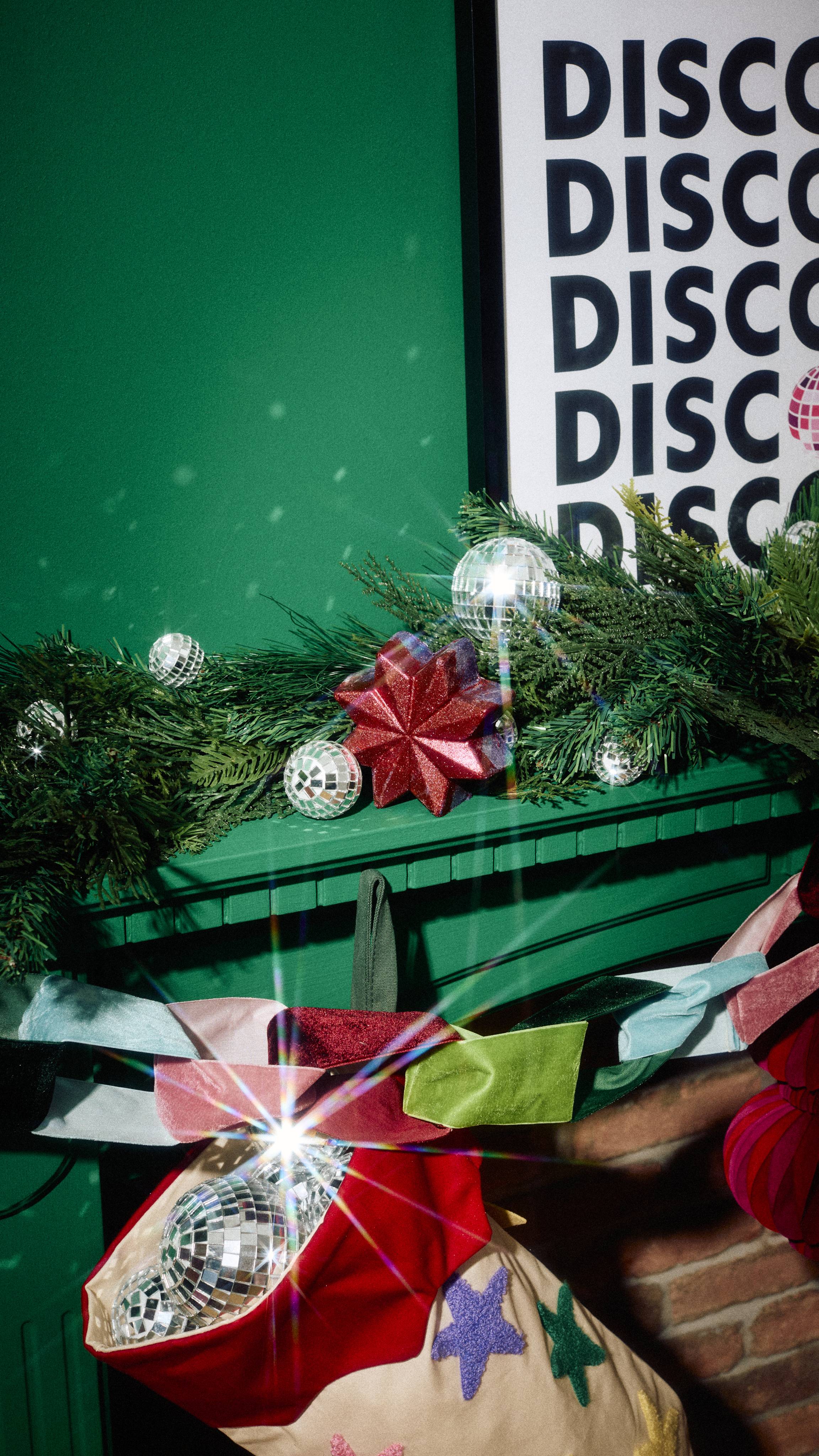 A shimmering ruby-red star bubble bar rests among miniature disco balls on a pine garland atop a green mantelpiece, decorated with velvet ribbon and a stocking filled with mirrored spheres.