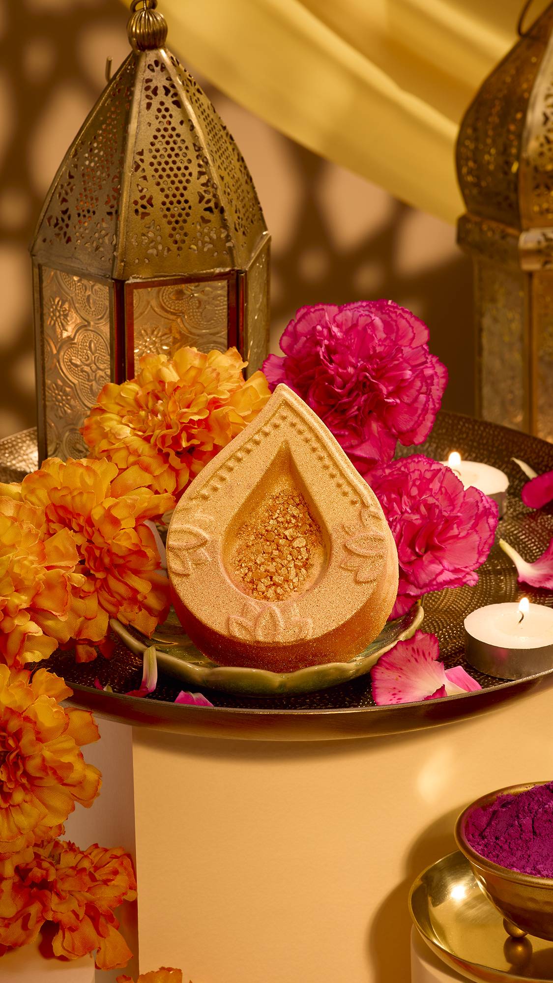 A golden, teardrop-shaped diya Lush bath bomb sits on a decorated tray with orange marigolds, magenta carnations, lit tealights, and ornate brass lanterns in a warm Diwali setting.