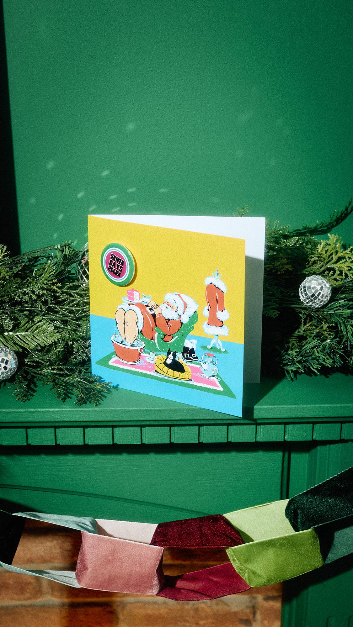 A Christmas card illustrating Santa relaxing in an armchair with a foot bath and cake, with an attached pin badge that says "Santa Says Relax" sits displayed on a green mantelpiece surrounded by pine garland, and festive decorations.