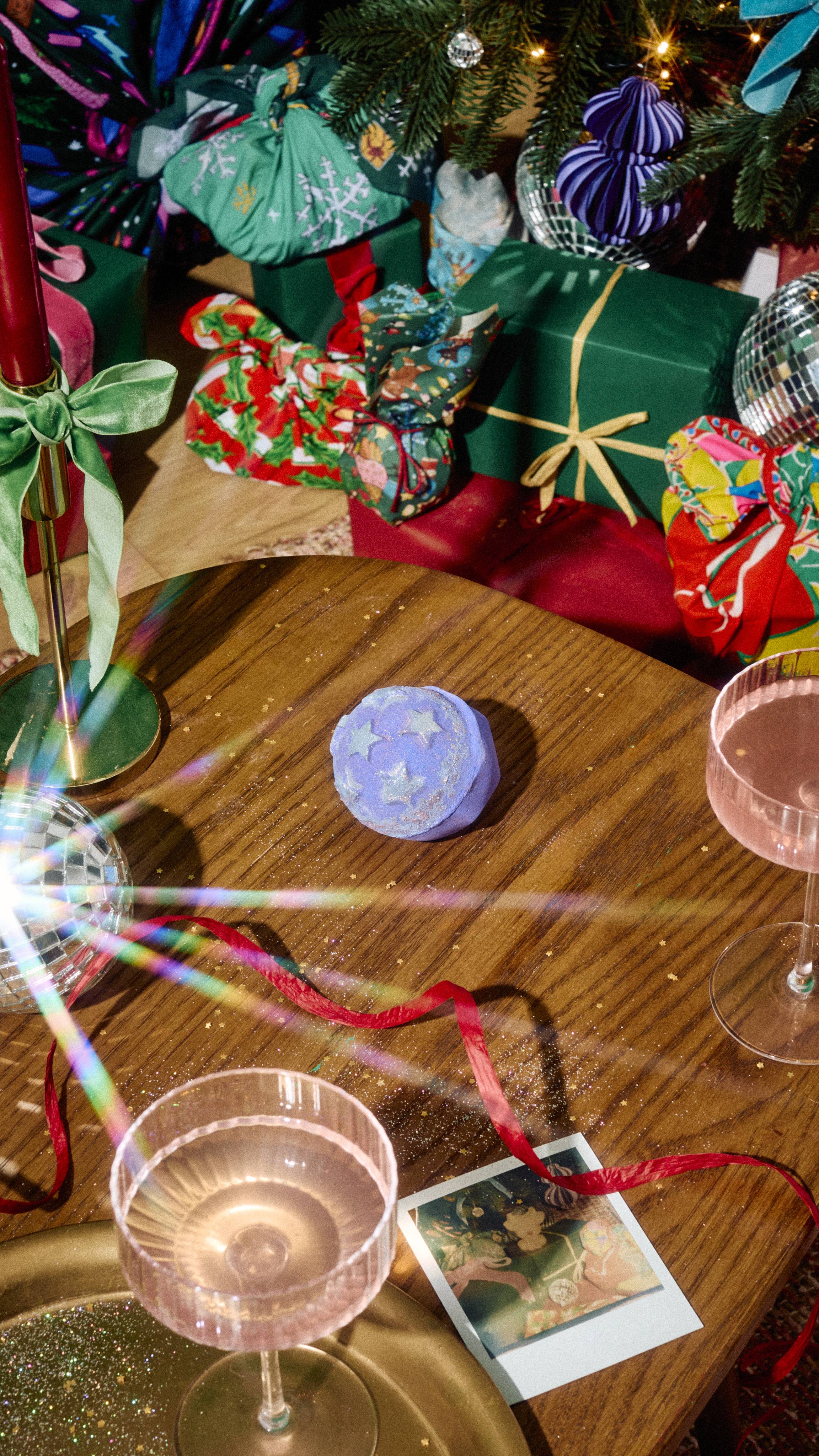 A spherical purple bath bomb embossed with light blue stars sits on a wooden table, surrounded by pink cocktail glasses, red ribbon, and wrapped Christmas gifts under a decorated tree.