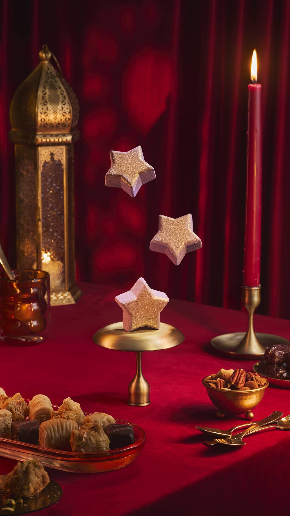 Three shimmering, star-shaped purple Siraj bath bombs appear to float above a small brass stand on a red velvet tablecloth. The festive scene includes an ornate gold lantern, a lit red candle, and bowls of nuts and pastries against red drapes.