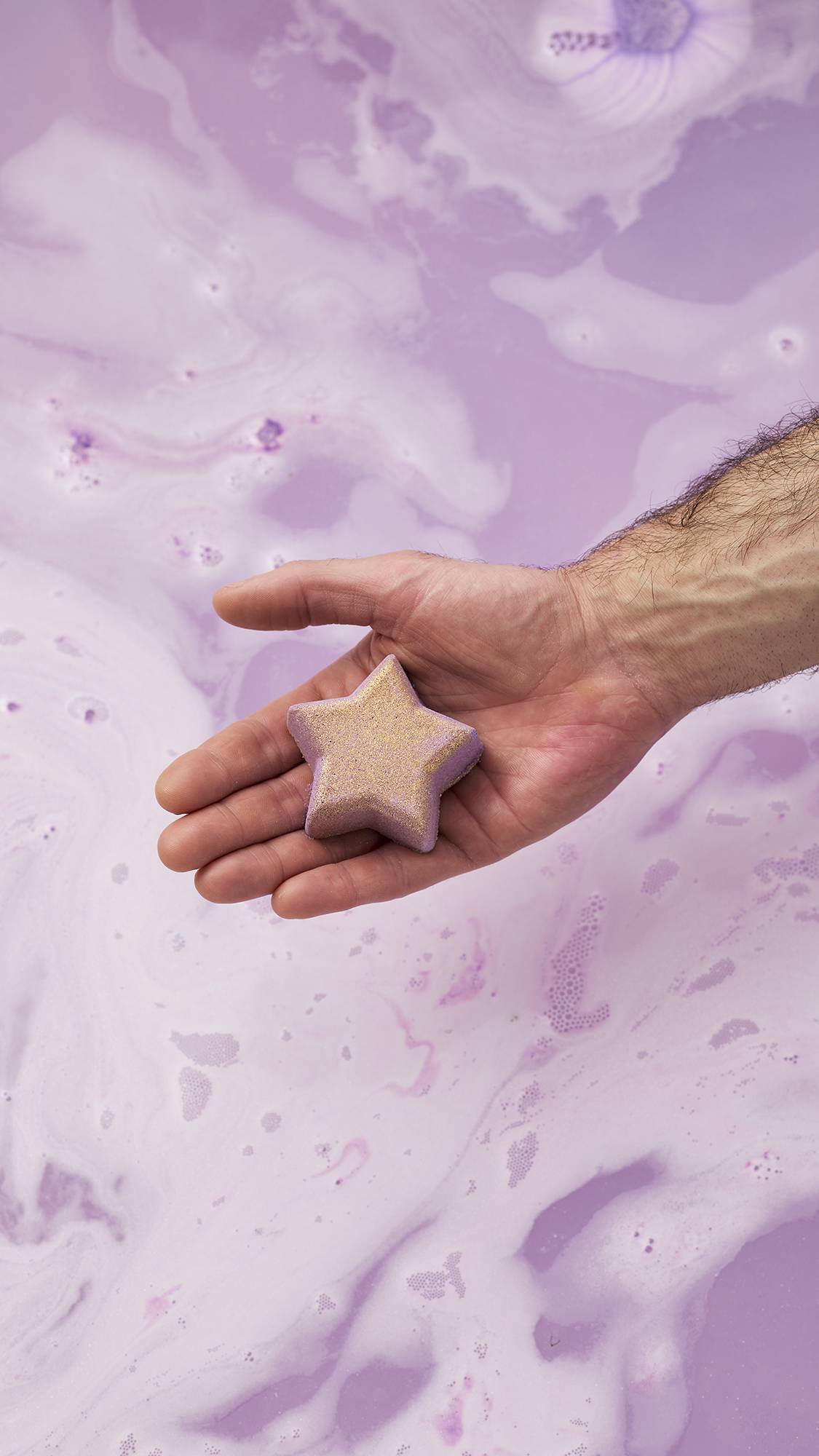 A person's open palm holds the gold-glittered, lilac star-shaped Siraj bath bomb over pale purple bath water filled with white foam and swirls.