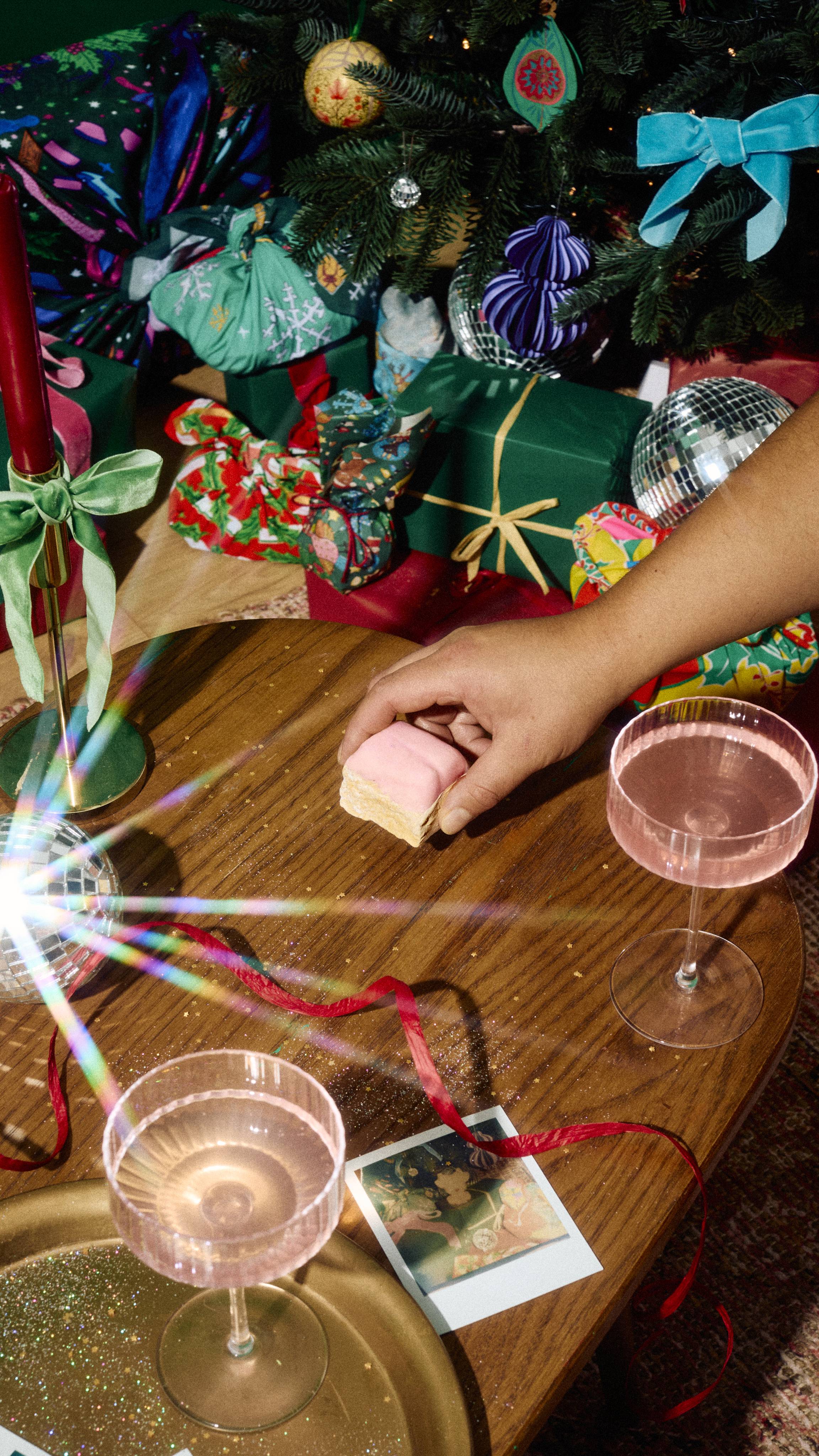 A hand reaches for a square "S'mores" soap with a pink top layer on a wooden table, surrounded by pink drinks, red ribbon, and wrapped gifts under a decorated Christmas tree.