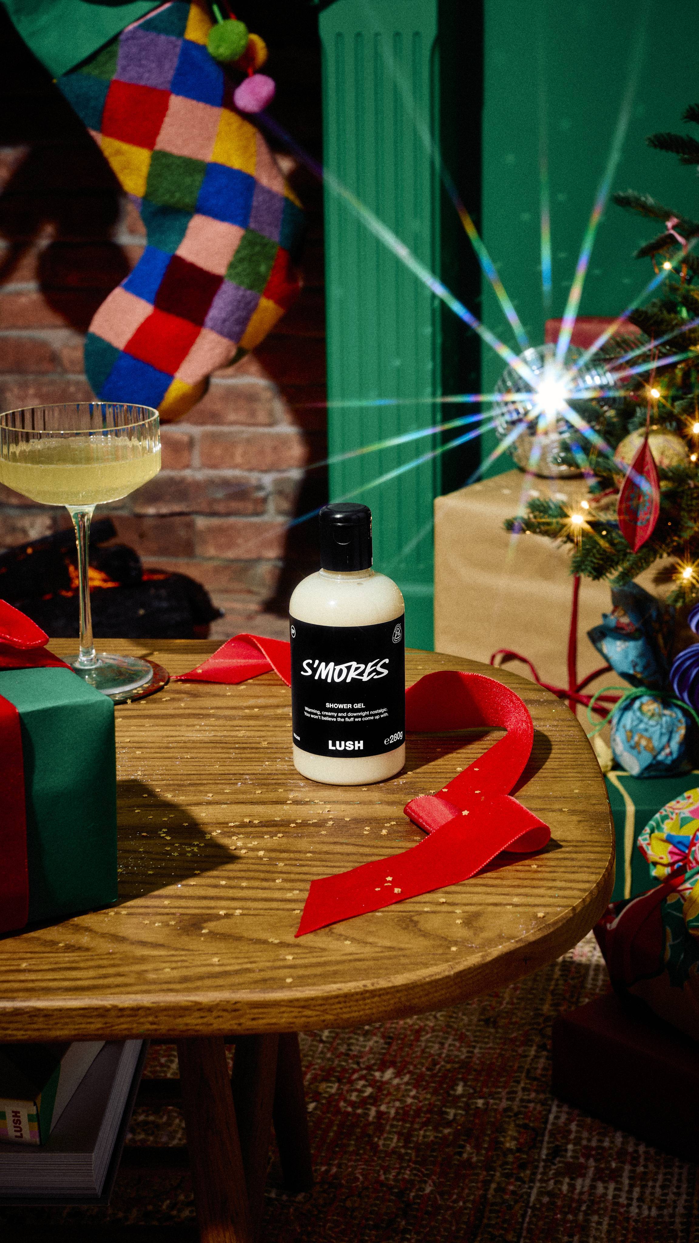 A bottle of Lush "S'mores" creamy white shower gel sits on a wooden table beside a glass of prosecco and red ribbon, with a colourful patchwork stocking and decorated Christmas tree in the background.