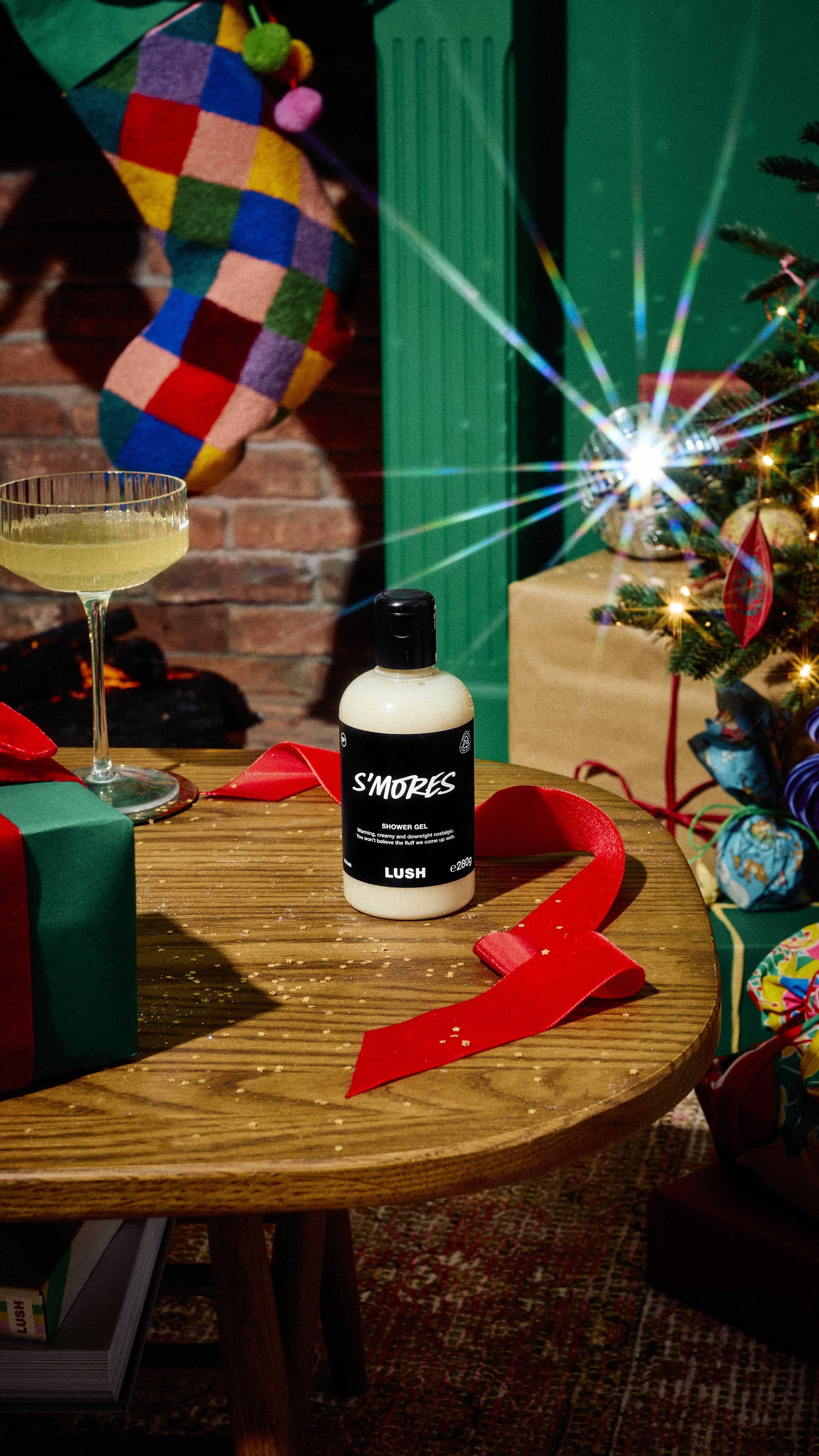 A bottle of Lush "S'mores" creamy white shower gel sits on a wooden table beside a glass of prosecco and red ribbon, with a colourful patchwork stocking and decorated Christmas tree in the background.