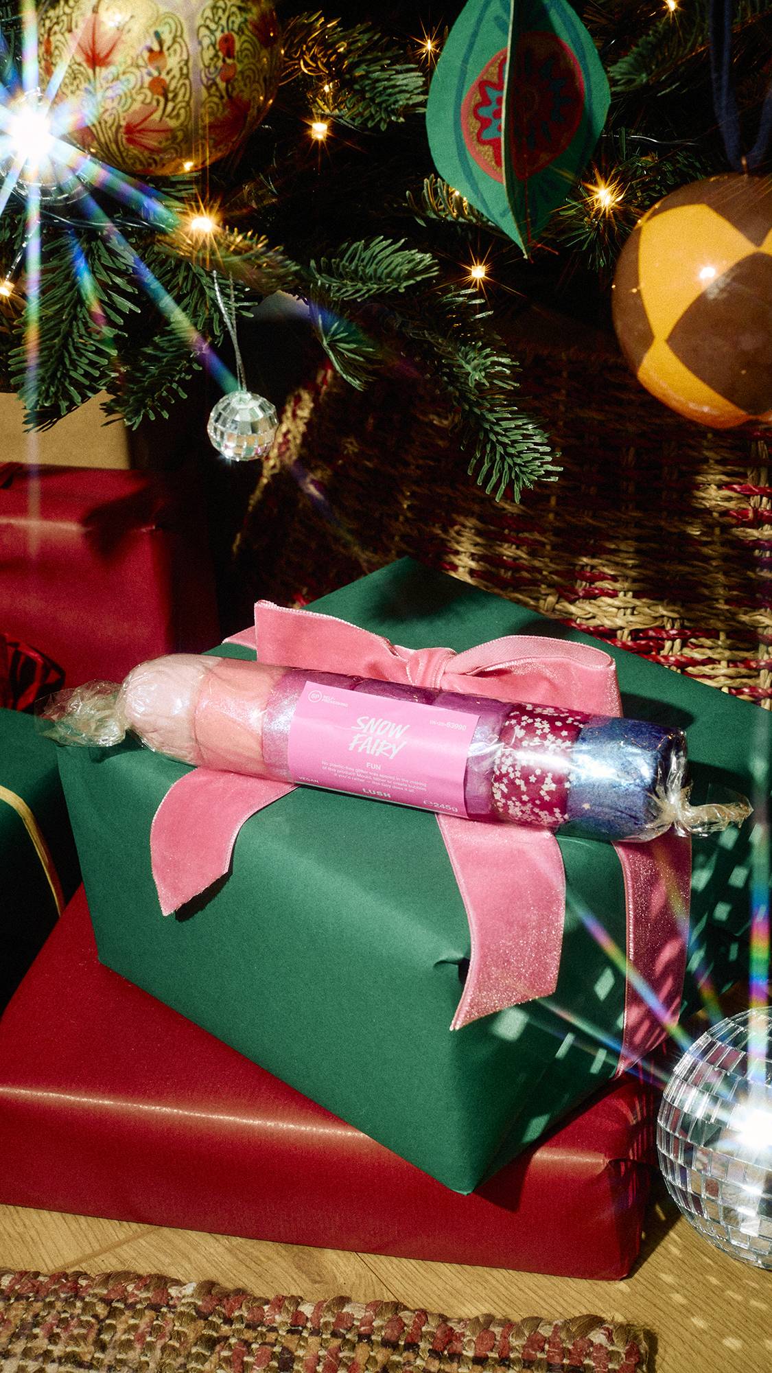 A clear, cracker-shaped wrapped Lush Snow Fairy FUN product with pink labelling rests on a green wrapped present tied with a pink velvet bow beneath a Christmas tree adorned with festive baubles.