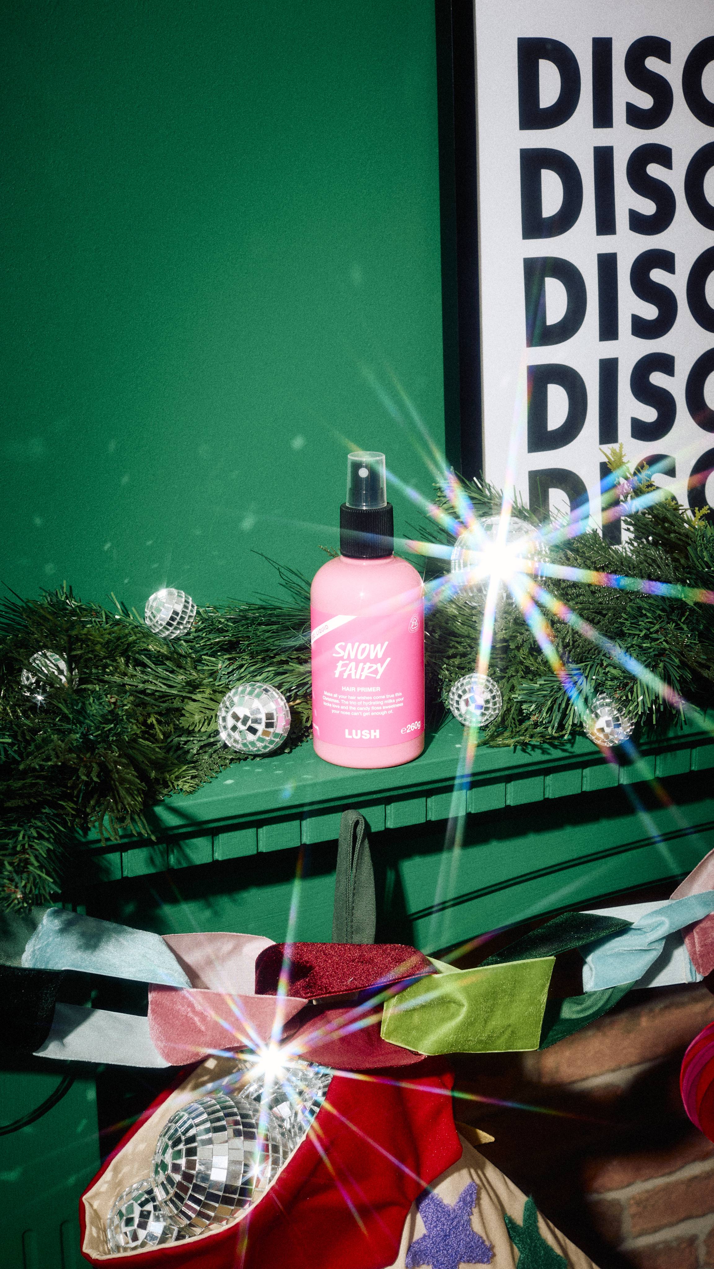 A bottle of Lush "Snow Fairy" pink hair primer spray stands on a green mantelpiece surrounded by pine garland, disco ball ornaments, and festive decorations against a green wall with framed typography art.