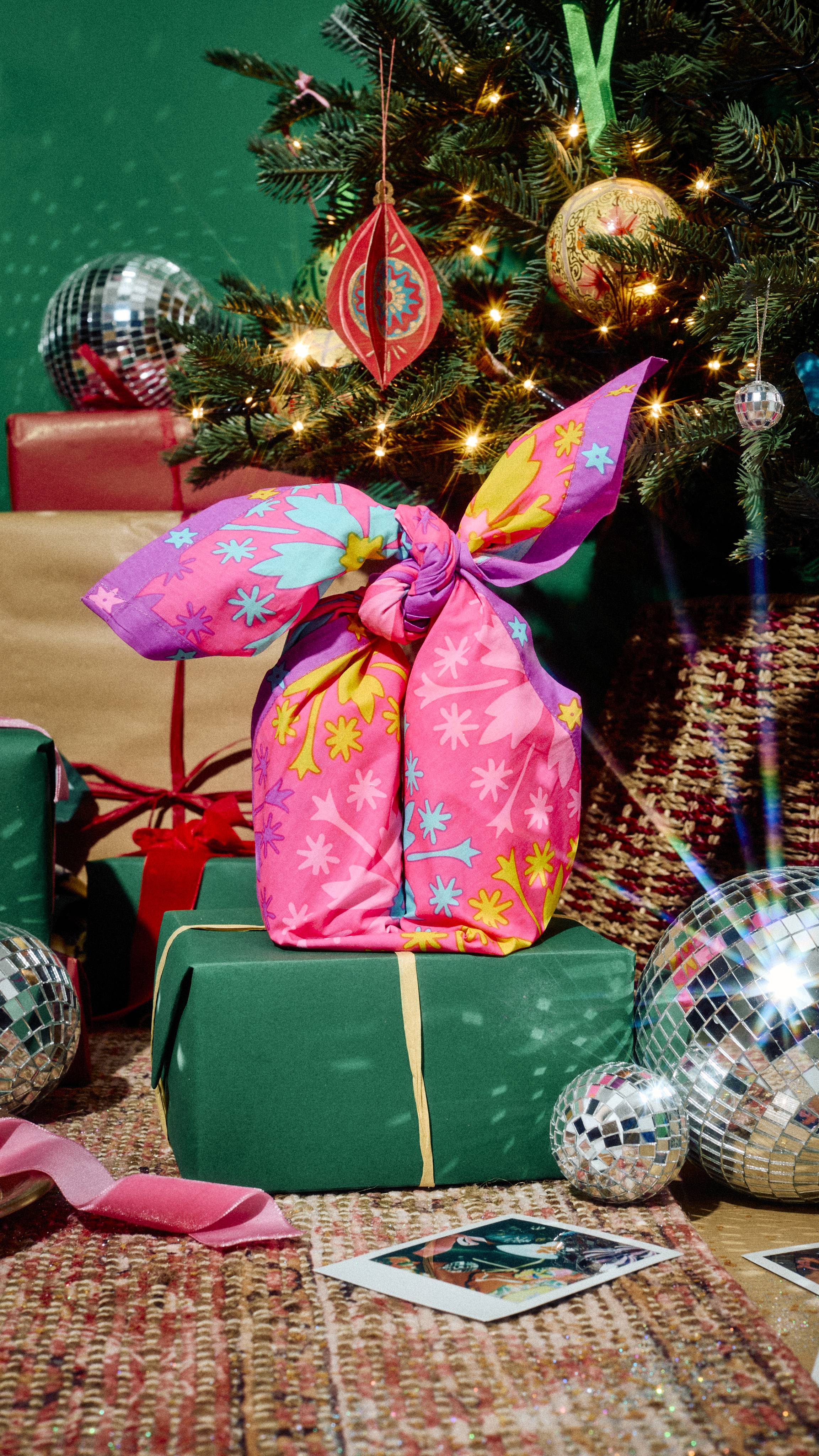 A gift wrapped in a vibrant pink floral Lush fabric "Snow Fairy" Knot Wrap sits atop a green gift box, placed beside a decorated Christmas tree with sparkly disco balls and other presents on a rug.