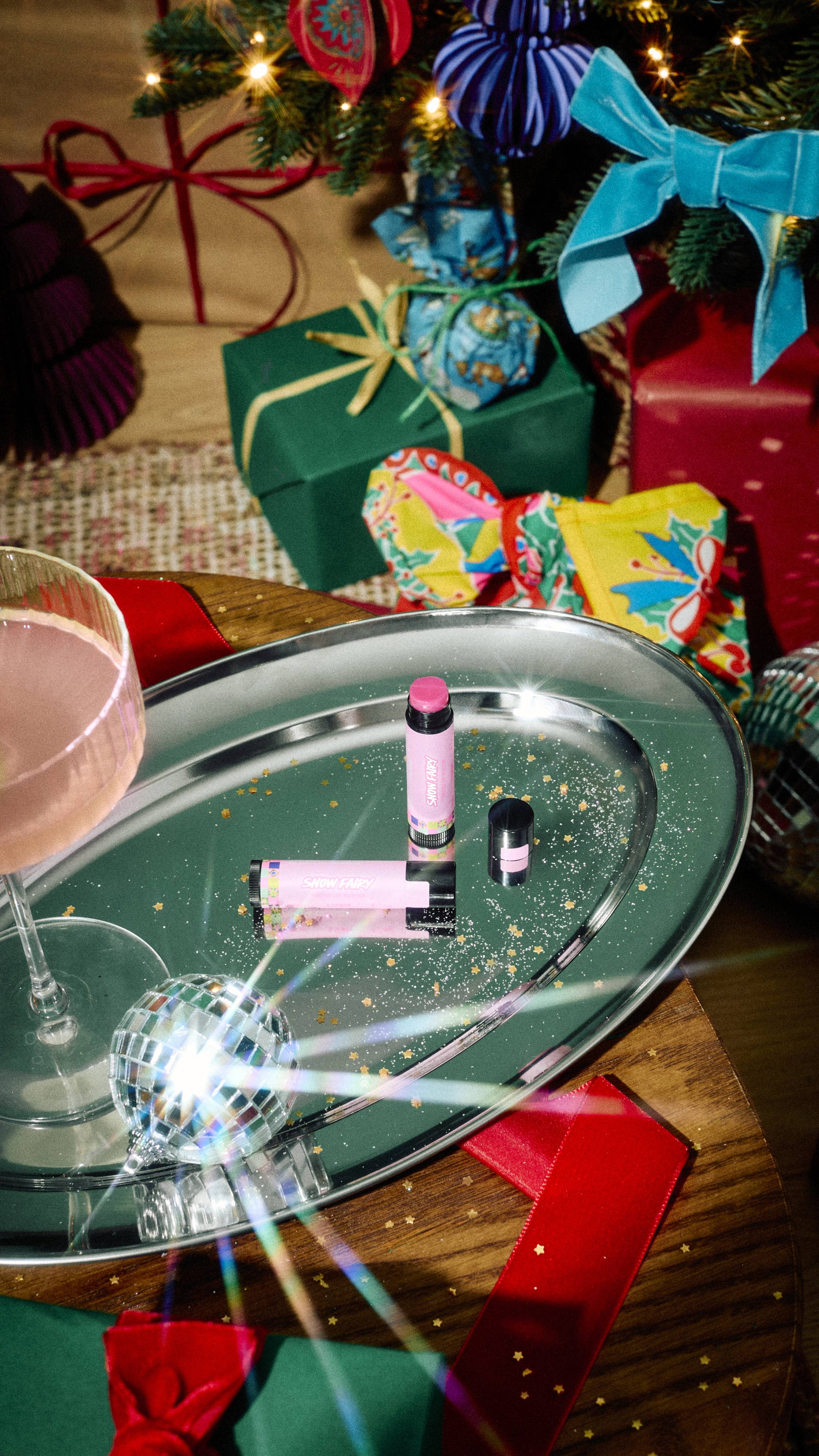 An open tube of pink Lush Snow Fairy tinted lip butter and its black cap rest on a silver tray with gold glitter, alongside a cocktail glass and mini disco ball, set against a festive backdrop of wrapped gifts.