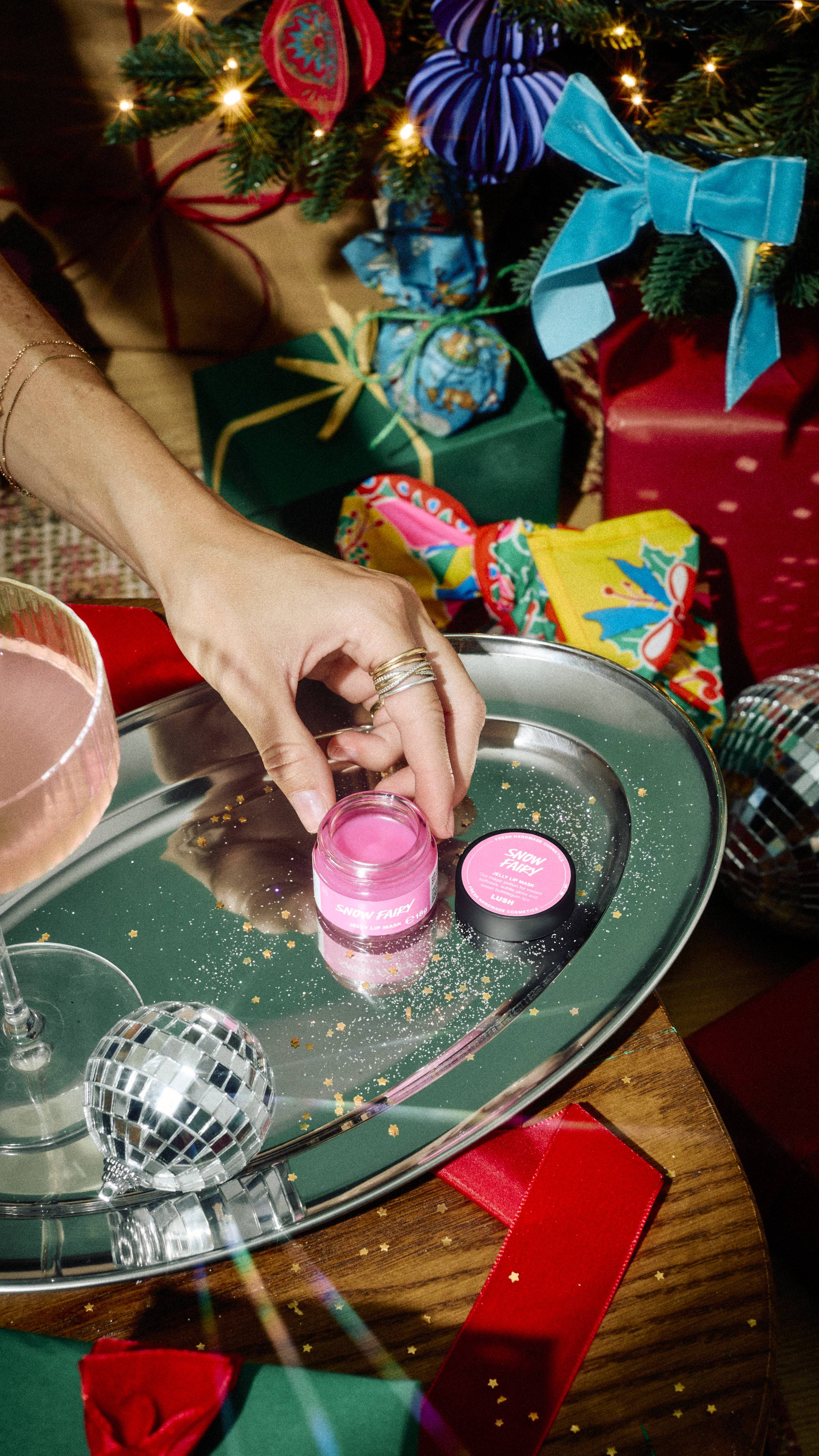 A hand reaches to place an open jar of pink Lush Snow Fairy lip product onto a silver tray alongside its black lid, set among festive gifts, a cocktail glass, and mini disco balls.