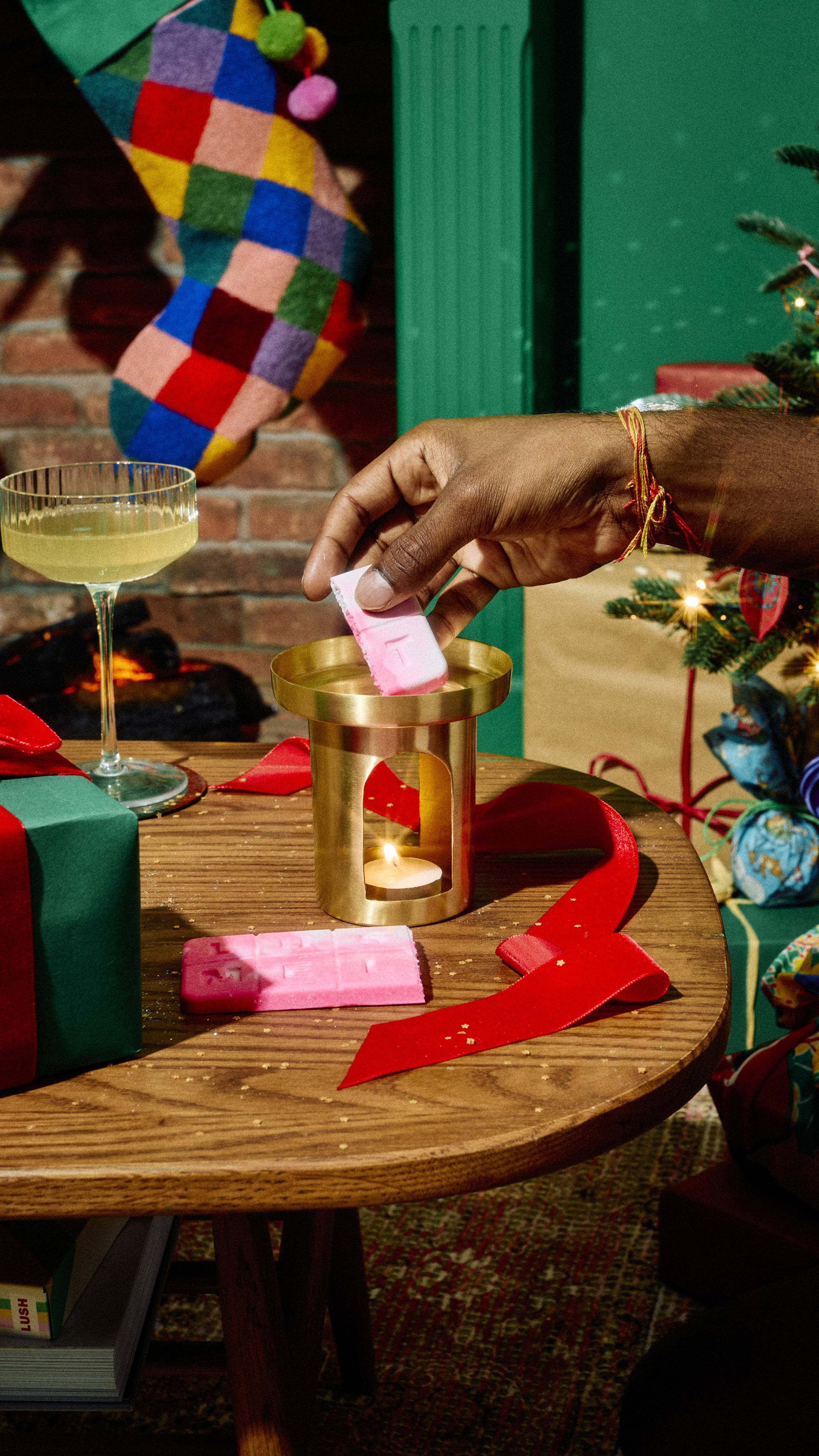 A hand places a piece of Lush "Snow Fairy" pink wax melt into a brass burner with a lit tealight inside, set on a wooden table with wrapped gifts, red ribbon, and a festive background.
