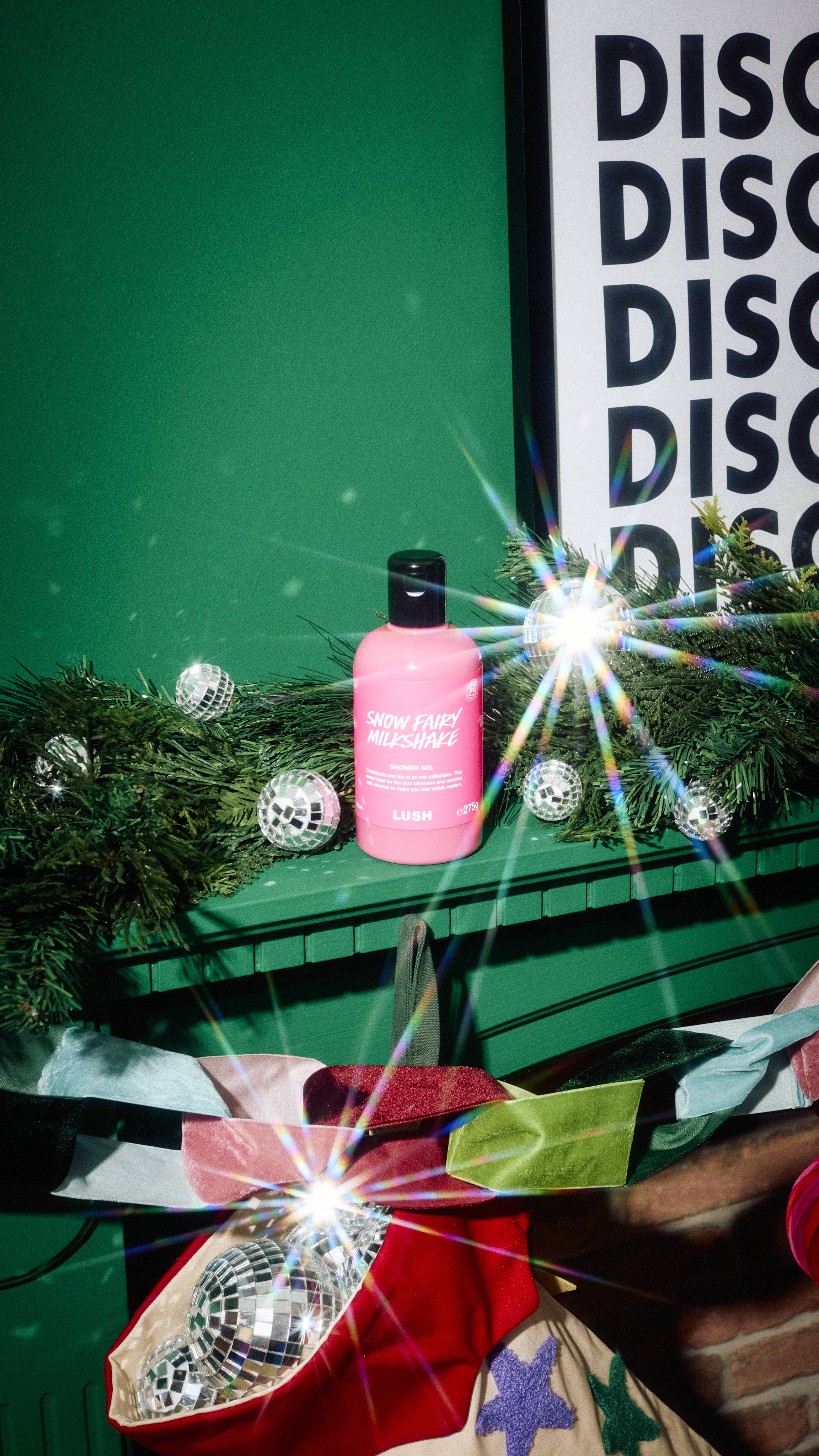 A bottle of Lush "Snow Fairy Milkshake" shower gel sits on a green mantelpiece alongside pine garland, mini disco balls, and a festive stocking, set against a dark green wall and framed "DISCO" print.