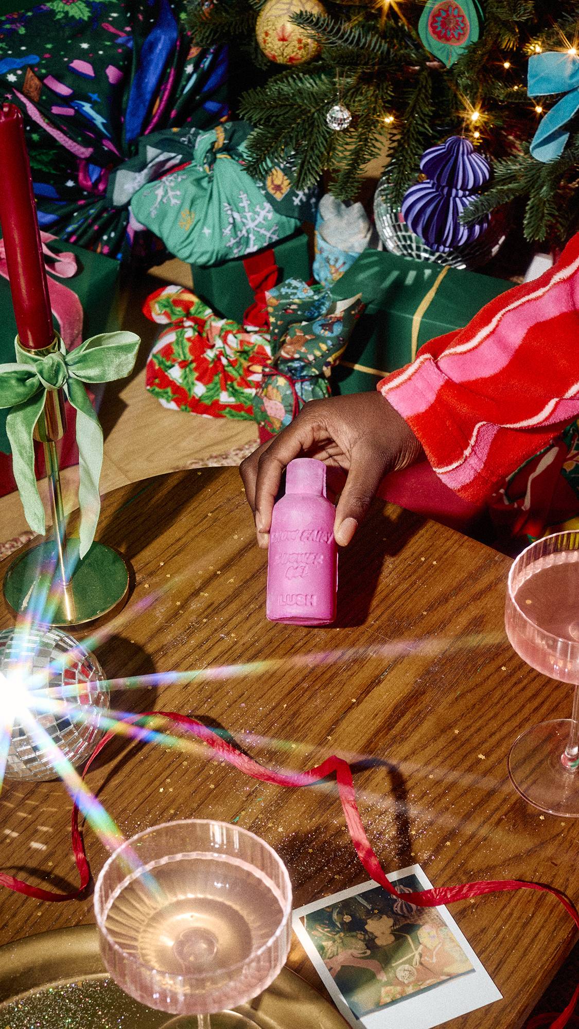 A hand places a bottle-shaped bar of pink Lush Snow Fairy naked shower gel on a wooden table styled with cocktail glasses, red ribbon, a tall candle, and mini disco balls beneath a decorated Christmas tree.