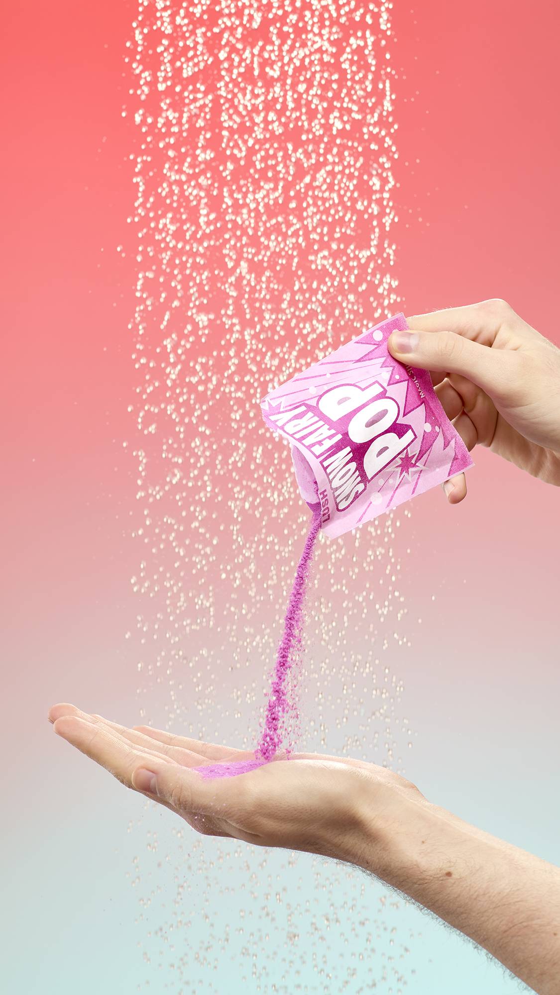 A person pours vibrant magenta Snow Fairy Pop shower powder from its pink sachet into their open palm. They are standing under a spray of falling water droplets against a soft pink-to-blue gradient background.
