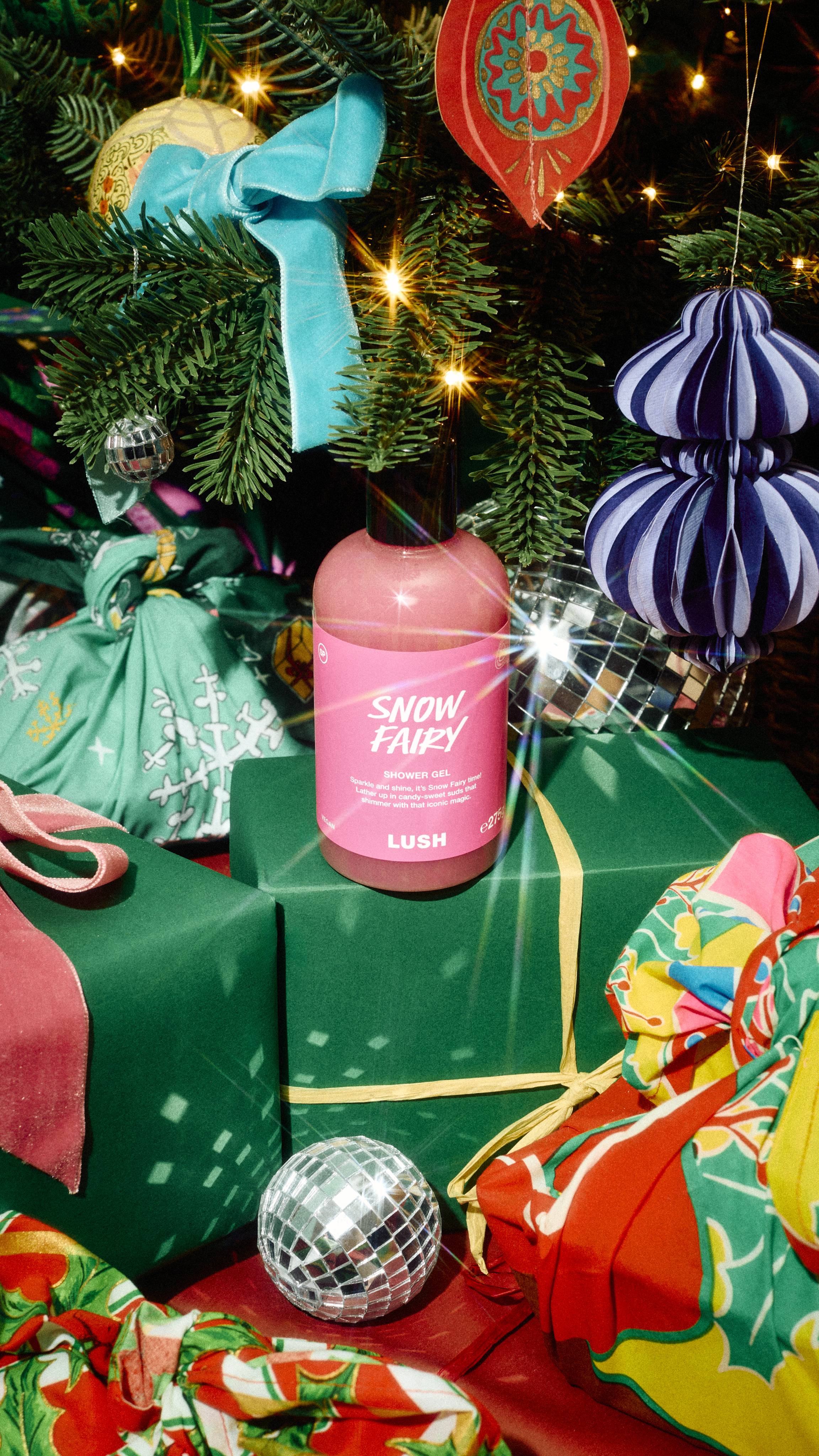 A bottle of Lush "Snow Fairy" pink shower gel sits on a green wrapped present tied with yellow ribbon beneath a decorated Christmas tree, surrounded by wrapped gifts, disco balls, and hanging ornaments.