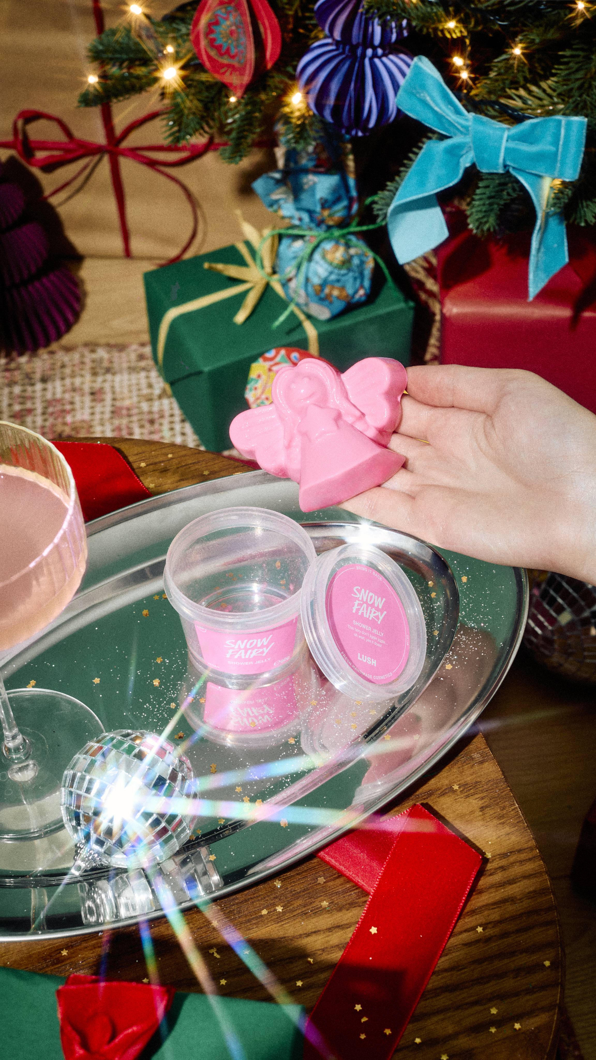 A hand holds a pink fairy-shaped shower jelly above a silver tray with a tub of Lush "Snow Fairy" product and a glass of pink drink, set against a festive Christmas background with wrapped gifts and tree decorations.