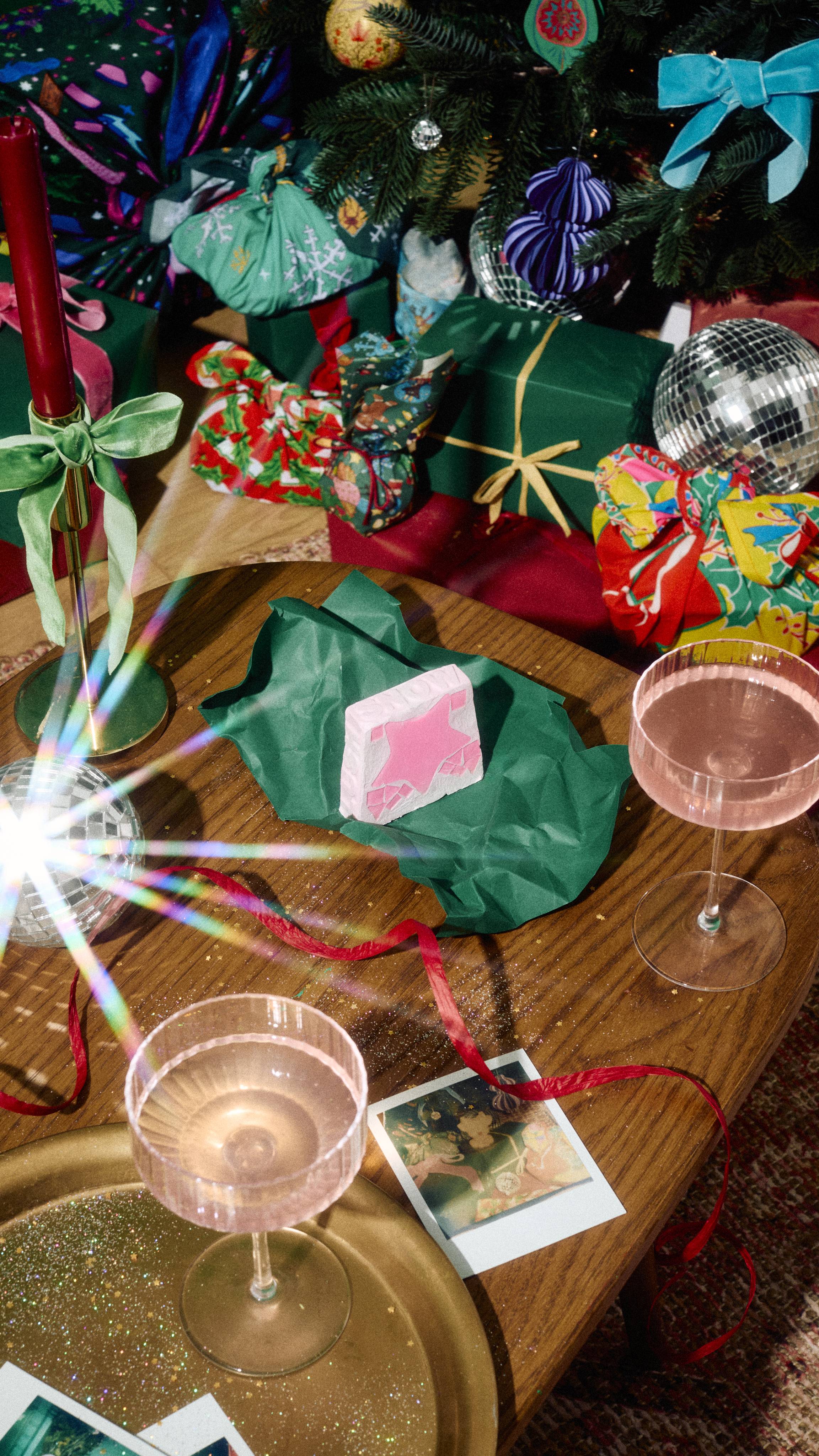 A pastel pink "Snow Fairy" soap featuring a dark pink star design rests on crumpled green tissue paper on a wooden table, flanked by pink drinks, red ribbon, and festive gifts under a Christmas tree.