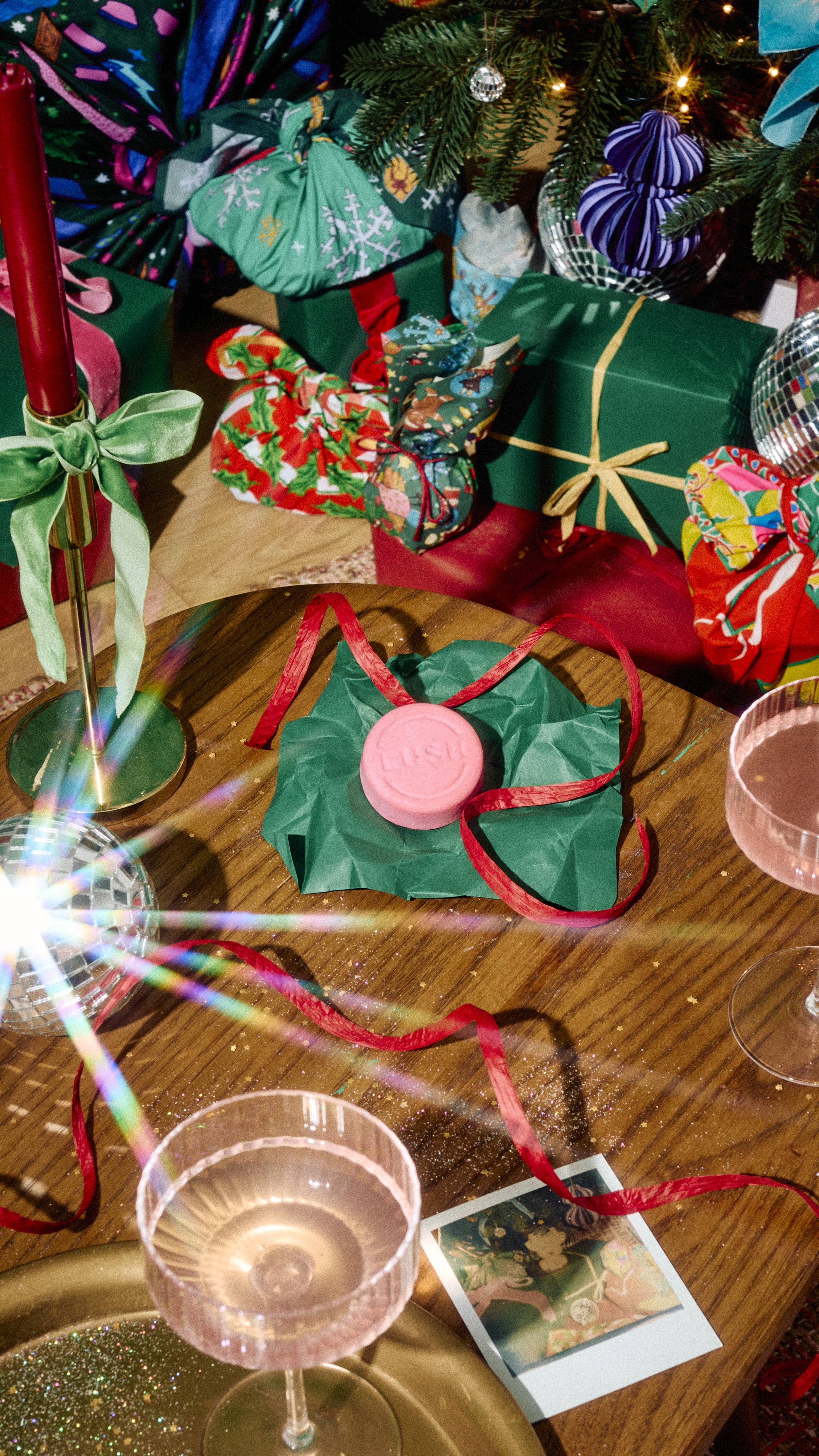 A round pink Lush "Snow Fairy" solid shampoo truckle bar sits on green tissue paper tied with red ribbon on a wooden table, surrounded by pink drinks and festive gifts under a decorated Christmas tree.