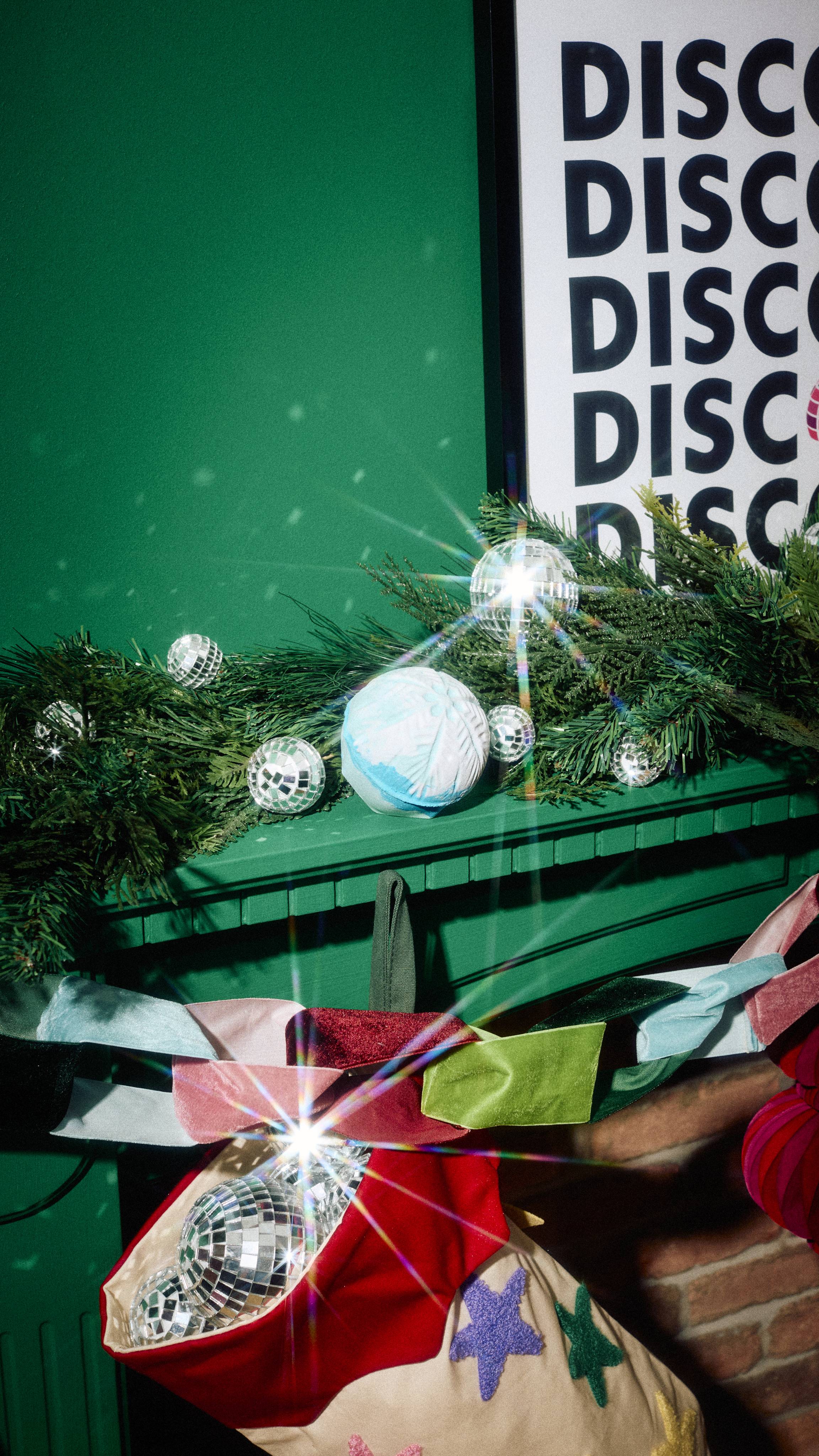 A spherical, light blue "Snowdrift" bath bomb rests on a green mantelpiece nestled in pine garland with mini disco balls, above a colourful velvet bow garland and a Christmas stocking filled with disco ball ornaments.