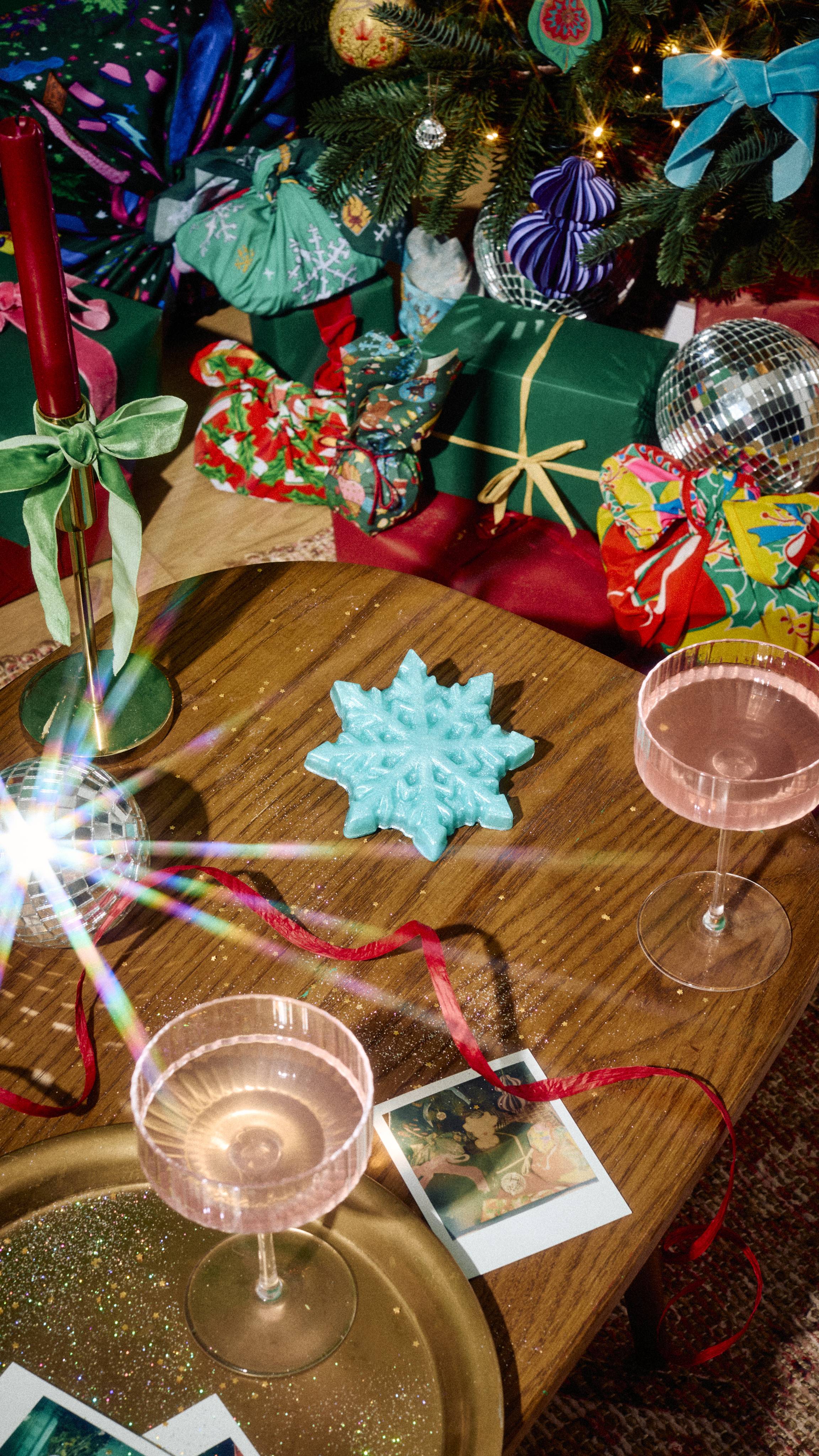 A shimmering mint green snowflake-shaped shower gummy sits on a wooden table alongside glasses of pink drink, red ribbon, and Polaroid photos, with wrapped Christmas presents beneath a decorated tree in the background.