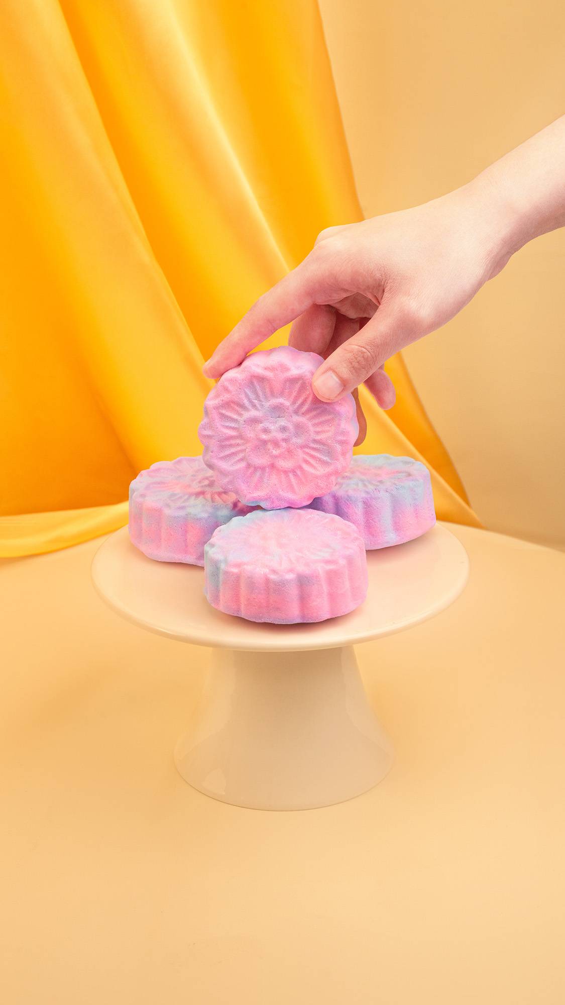 A hand places a pink and blue marbled, mooncake-shaped bubble bars onto a small stack of identical items on a cream cake stand. The arrangement is set against a warm beige background with a draped gold fabric backdrop.