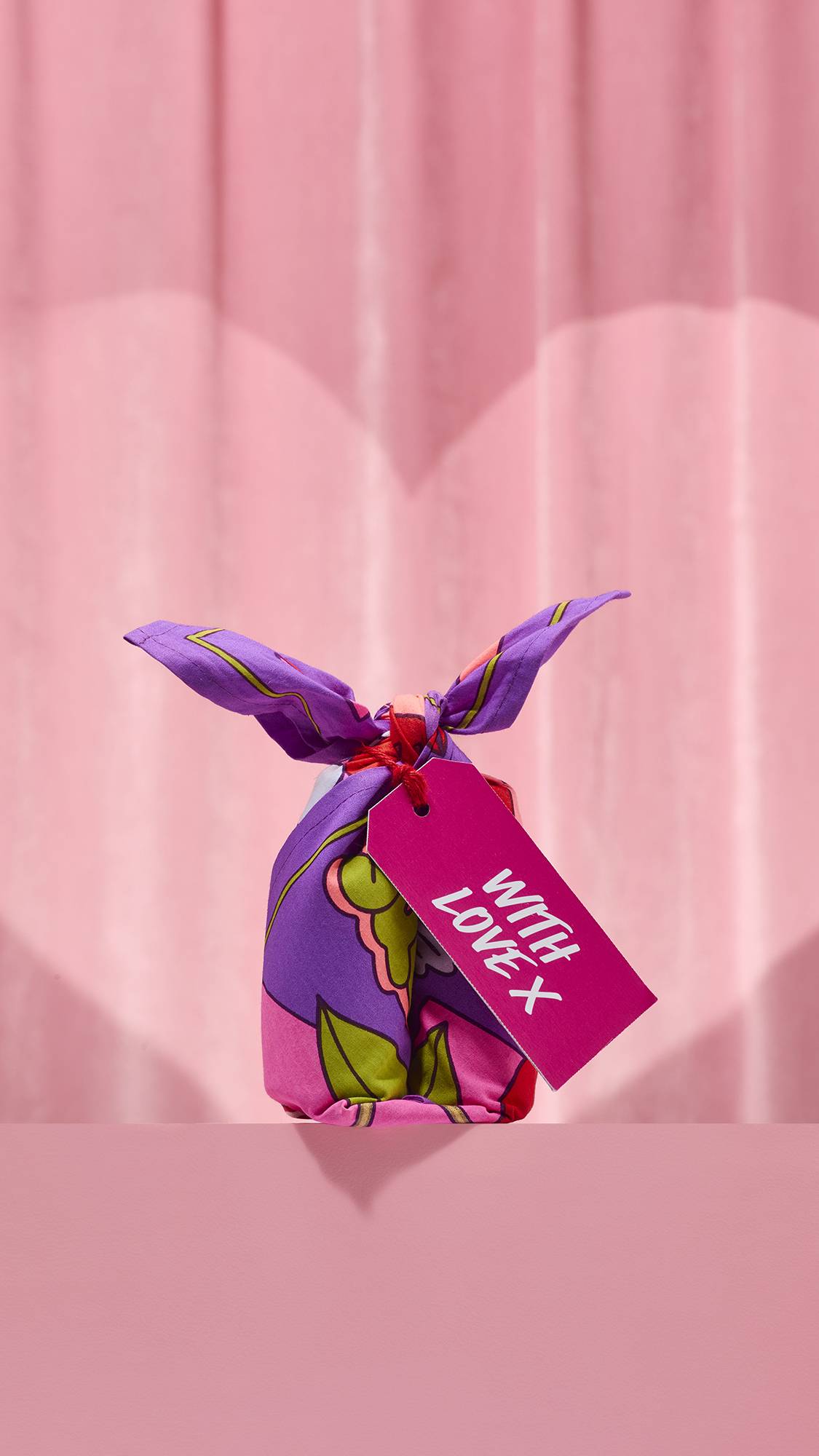 The Squeeze Knot Wrap features purple fabric with a pattern of green leaves and yellow accents tied around a small gift. A hot pink tag reading "WITH LOVE X" hangs from the knot, set against a soft pink curtain background.