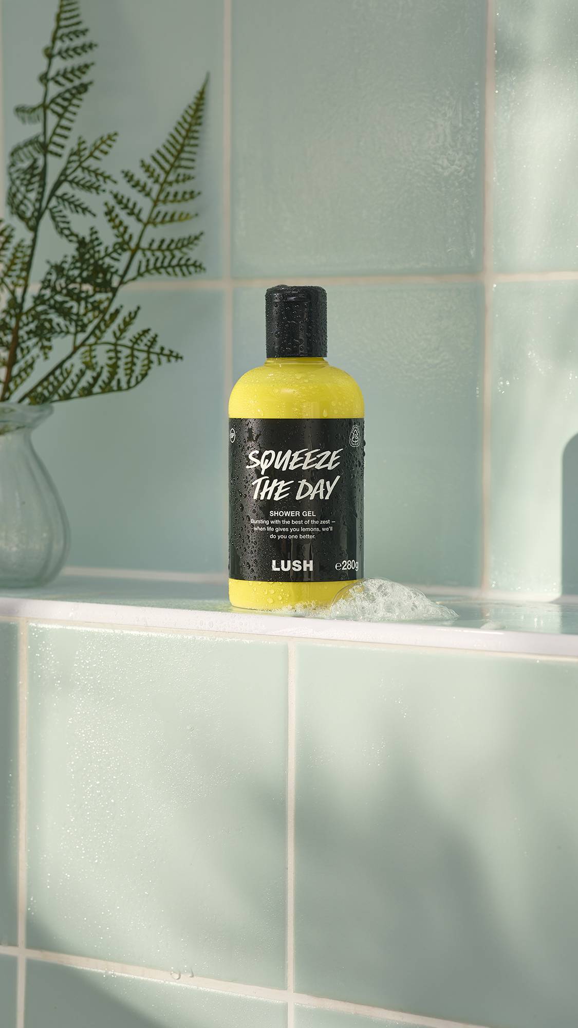 A bottle of yellow "Squeeze The Day" shower gel by Lush sits on a pale green tiled ledge. The bottle is covered in water droplets and has a small patch of white foam at its base. A green fern in a vase is partially visible in the background.
