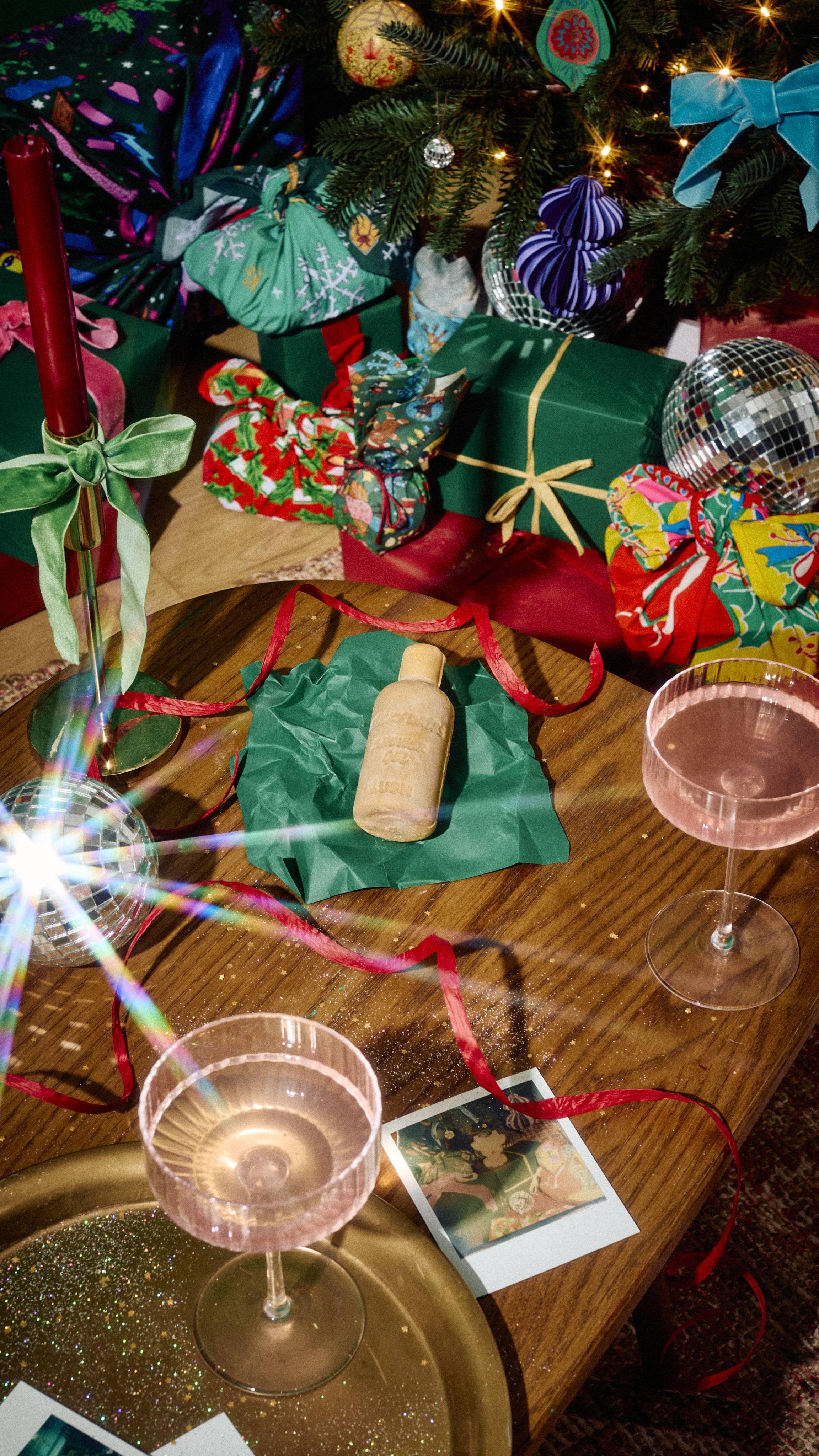 A bottle-shaped bar of beige Lush Sticky Dates naked shower gel rests on green tissue paper on a wooden table, surrounded by cocktail glasses, red ribbon, and mini disco balls under a decorated Christmas tree.