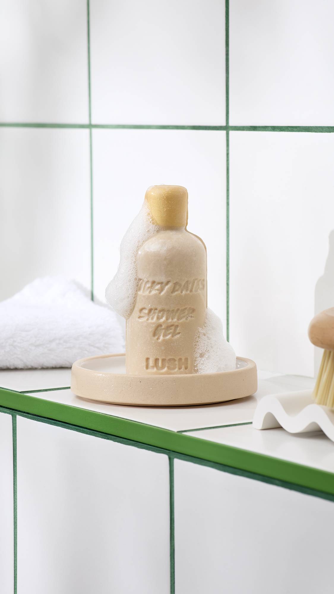 A bottle-shaped bar of beige Lush Sticky Dates naked shower gel, covered in white foamy lather, rests on a small ceramic dish in a shower setting with white tiled walls and a folded white towel.