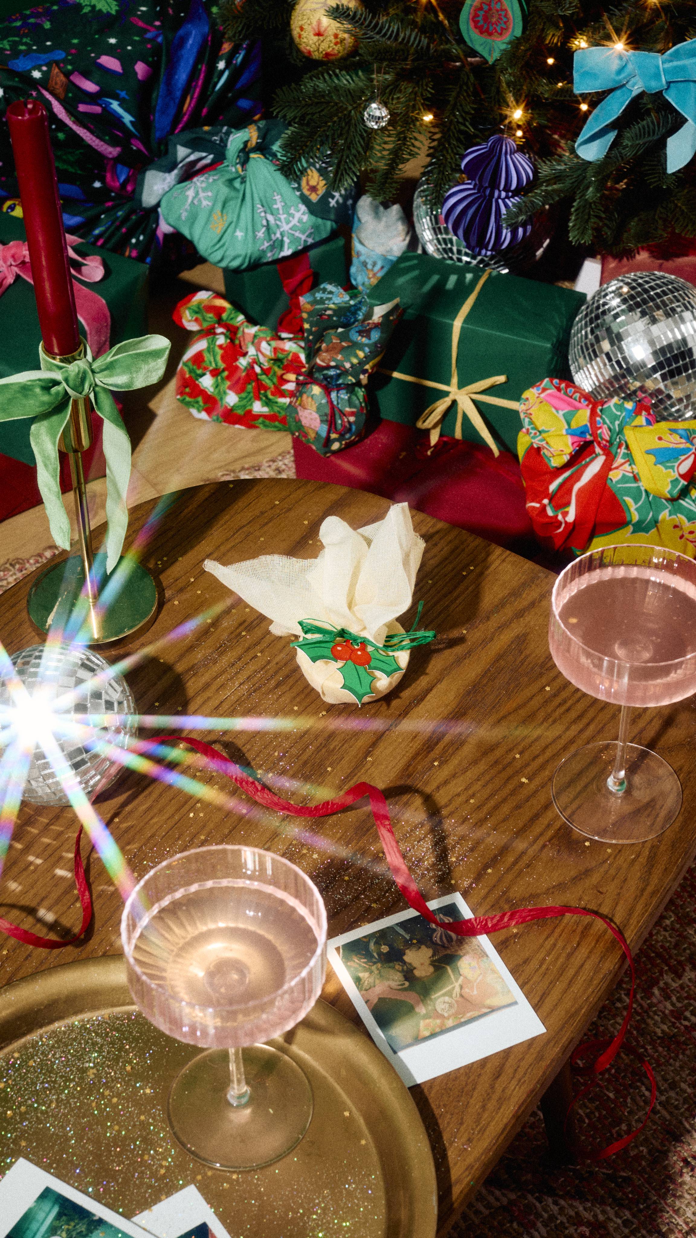 A Lush "Sticky Toffee" bath melt wrapped in mesh fabric with a holly tag sits on a wooden table alongside glasses of pink drink, red ribbon, and wrapped gifts under a decorated Christmas tree.