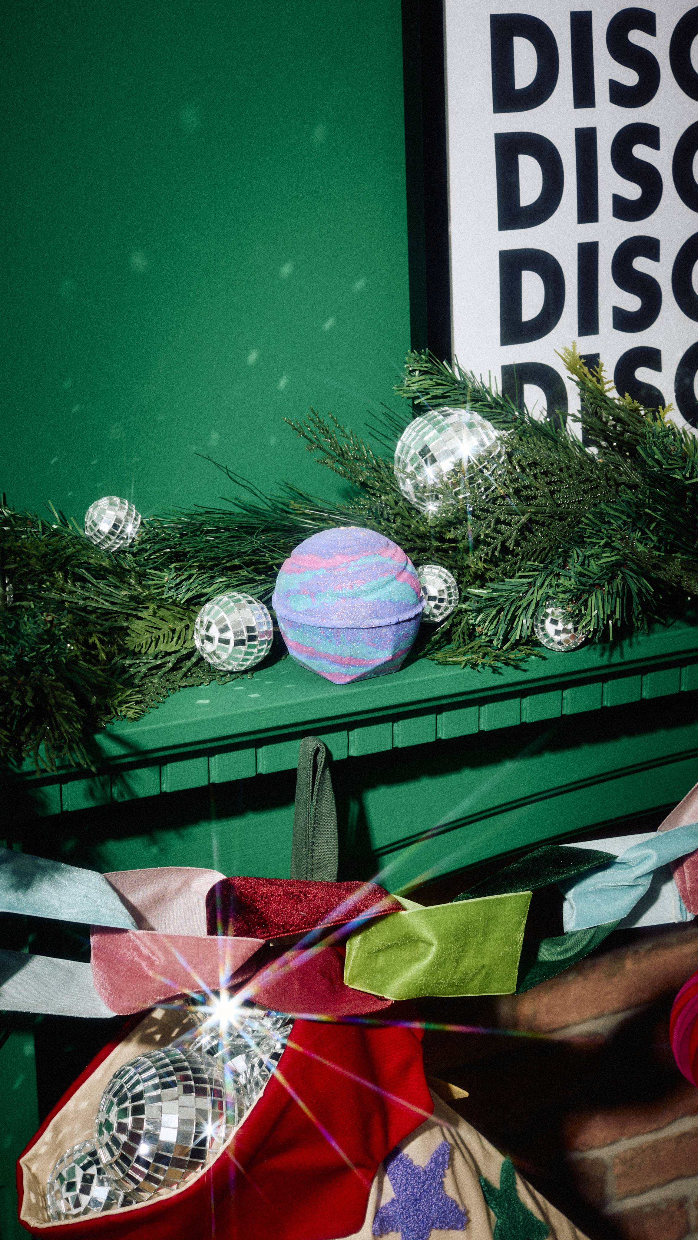 A round bath bomb with striped purple, pink, and blue hues rests on a green mantelpiece surrounded by pine garland, mini disco balls, a colourful velvet bow garland, and a stocking stuffed with shiny disco ball ornaments.