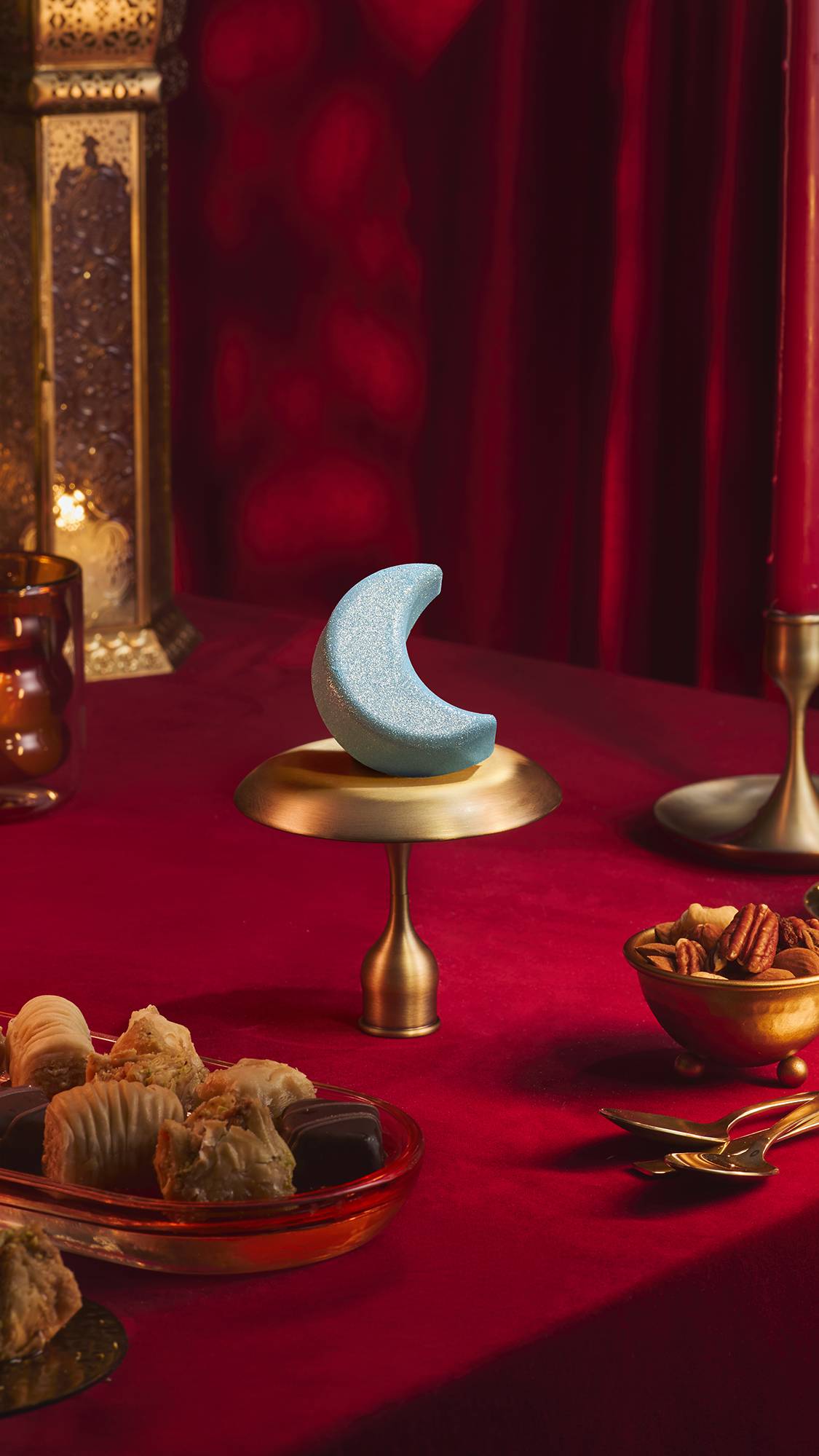 A blue, shimmering, crescent moon-shaped Sukoon bath bomb sits on a small brass stand atop a red velvet tablecloth. Surrounding it are bowls of nuts, pastries, and an ornate golden lantern against a backdrop of draped red fabric.